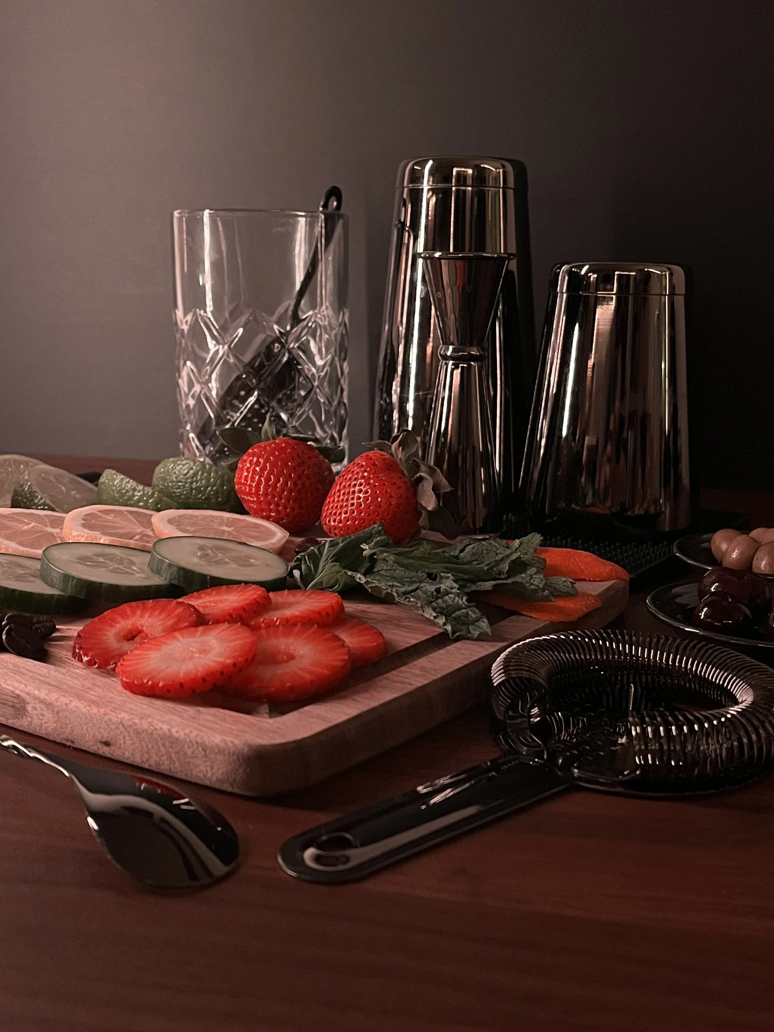 A bar with sliced strawberries, lemons, and oranges on a wooden cutting board, a clear glass, and cocktail shakers on a dark background.