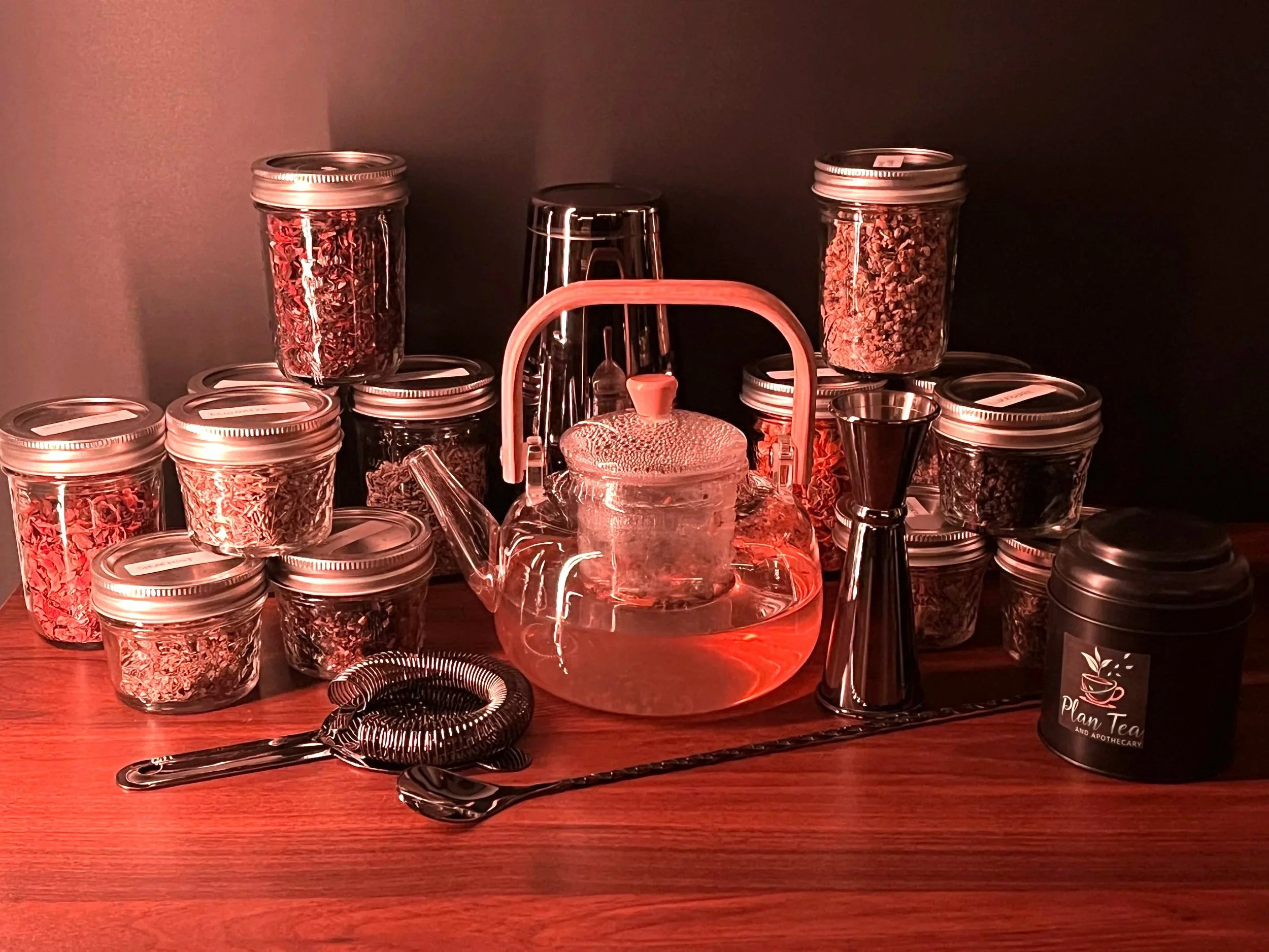 A variety of glass jars filled with dried herbs or tea leaves, a glass teapot, tea infuser, and a black tin labeled 'Plan Tea' on a wooden table, illuminated by red lighting.