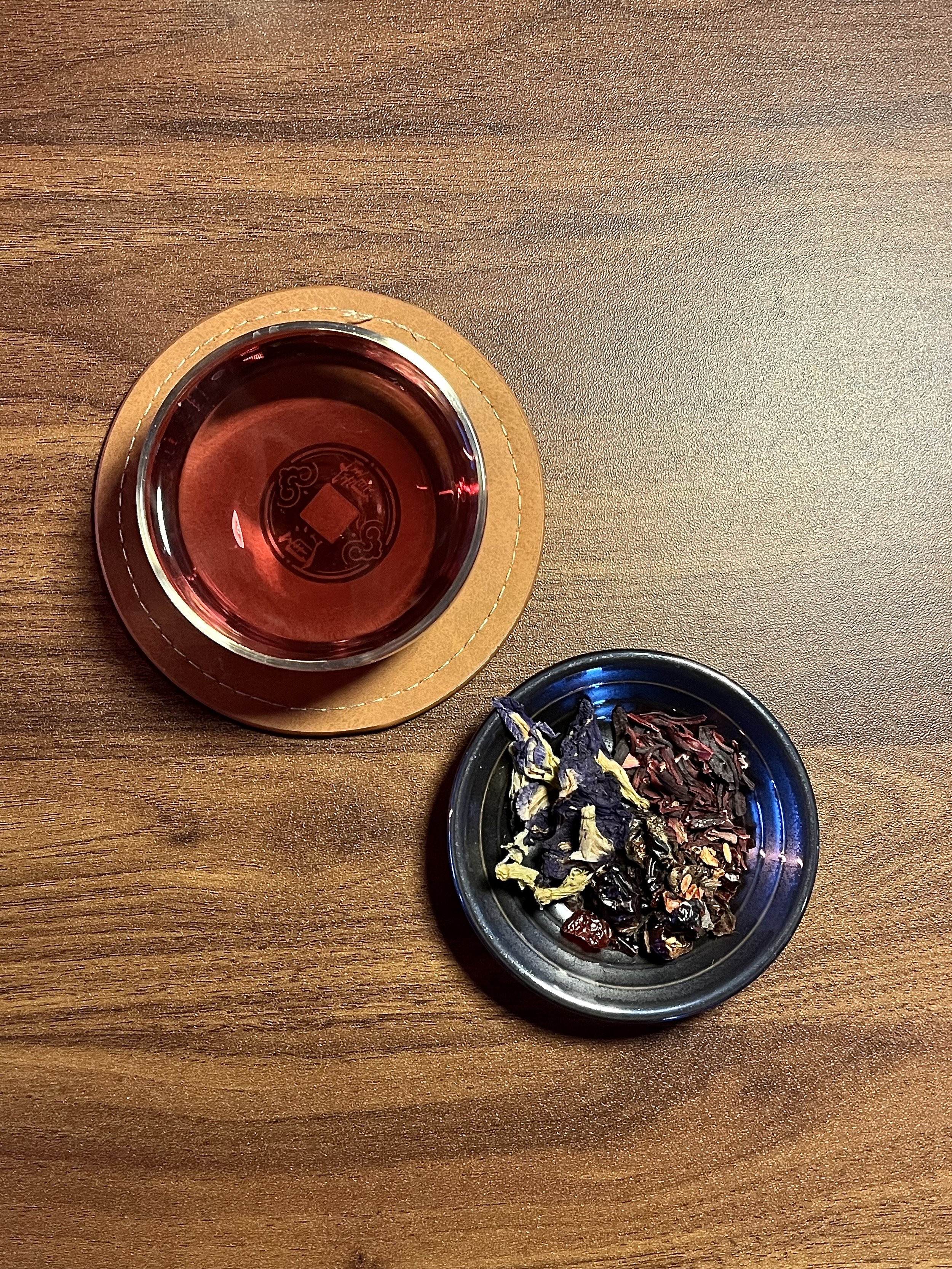 A wooden table with a small glass of red tea on a circular coaster and a small dish of dried herbs or flowers.