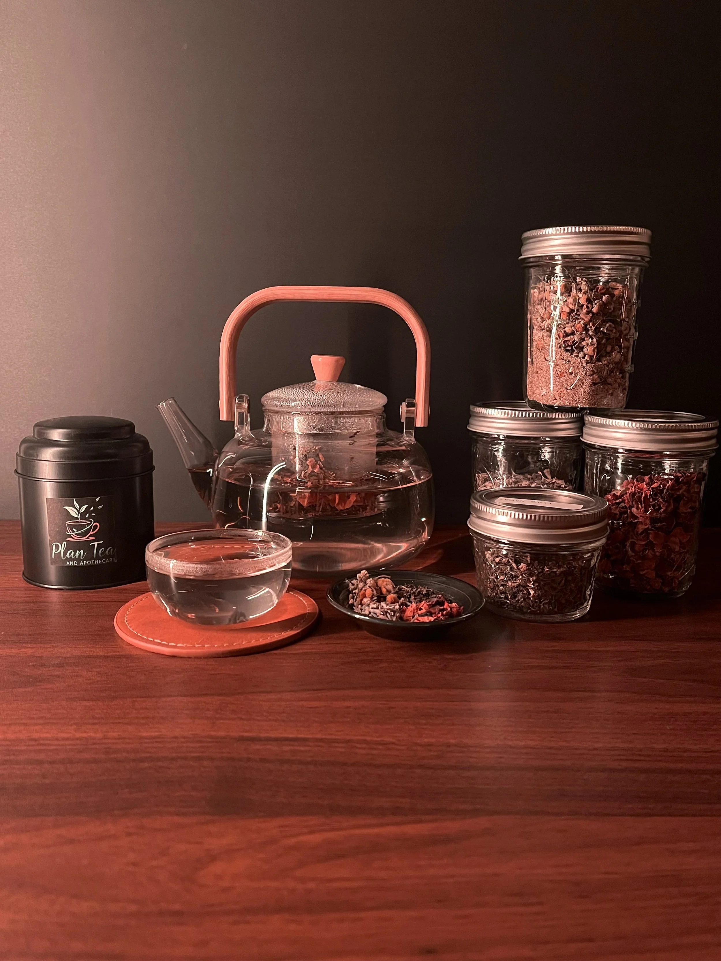 A glass teapot filled with water, sealed with a pink handle, surrounded by various jars and containers of dried herbs and tea leaves on a wooden table against a dark background.