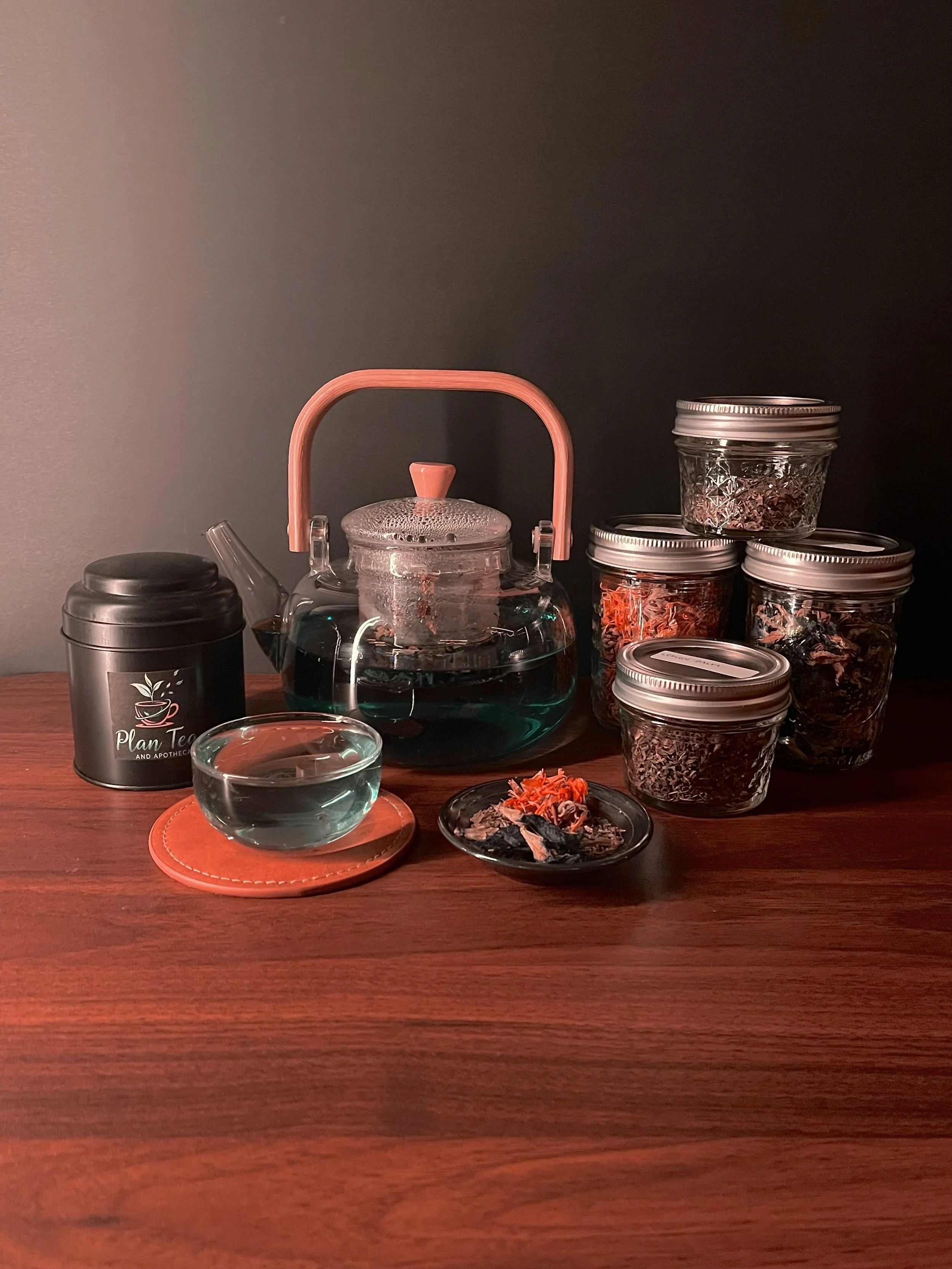 A wooden table with a glass teapot filled with tea, a glass cup on a coaster, a black container with a planning logo, and multiple glass jars filled with dried flowers and herbs. The background is a dark wall.