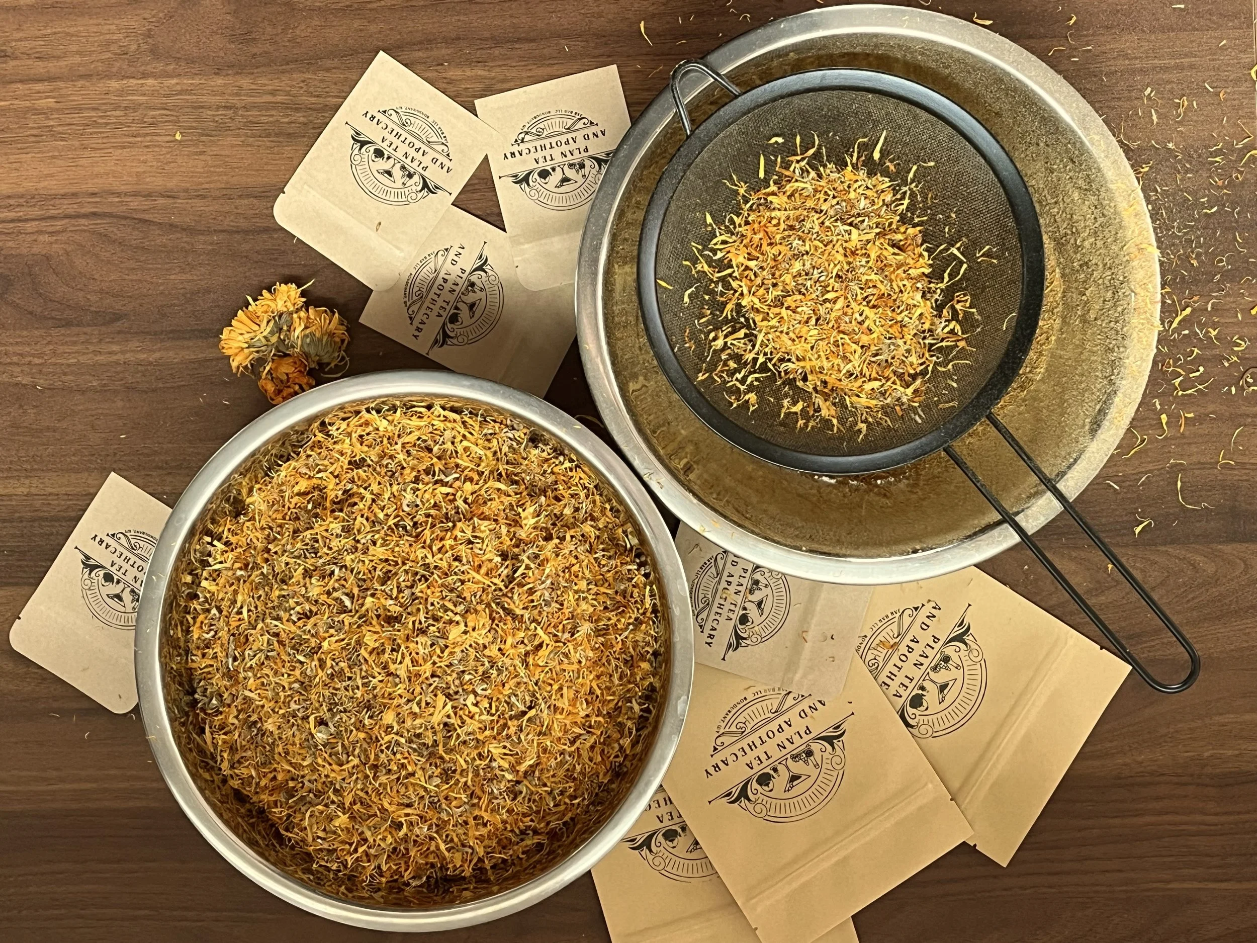 Dried calendula flower petals being sifted in a metal strainer on a wooden surface, with paper bags labeled "Plant Tea and Apothecary" and a small dried flower bundle nearby.