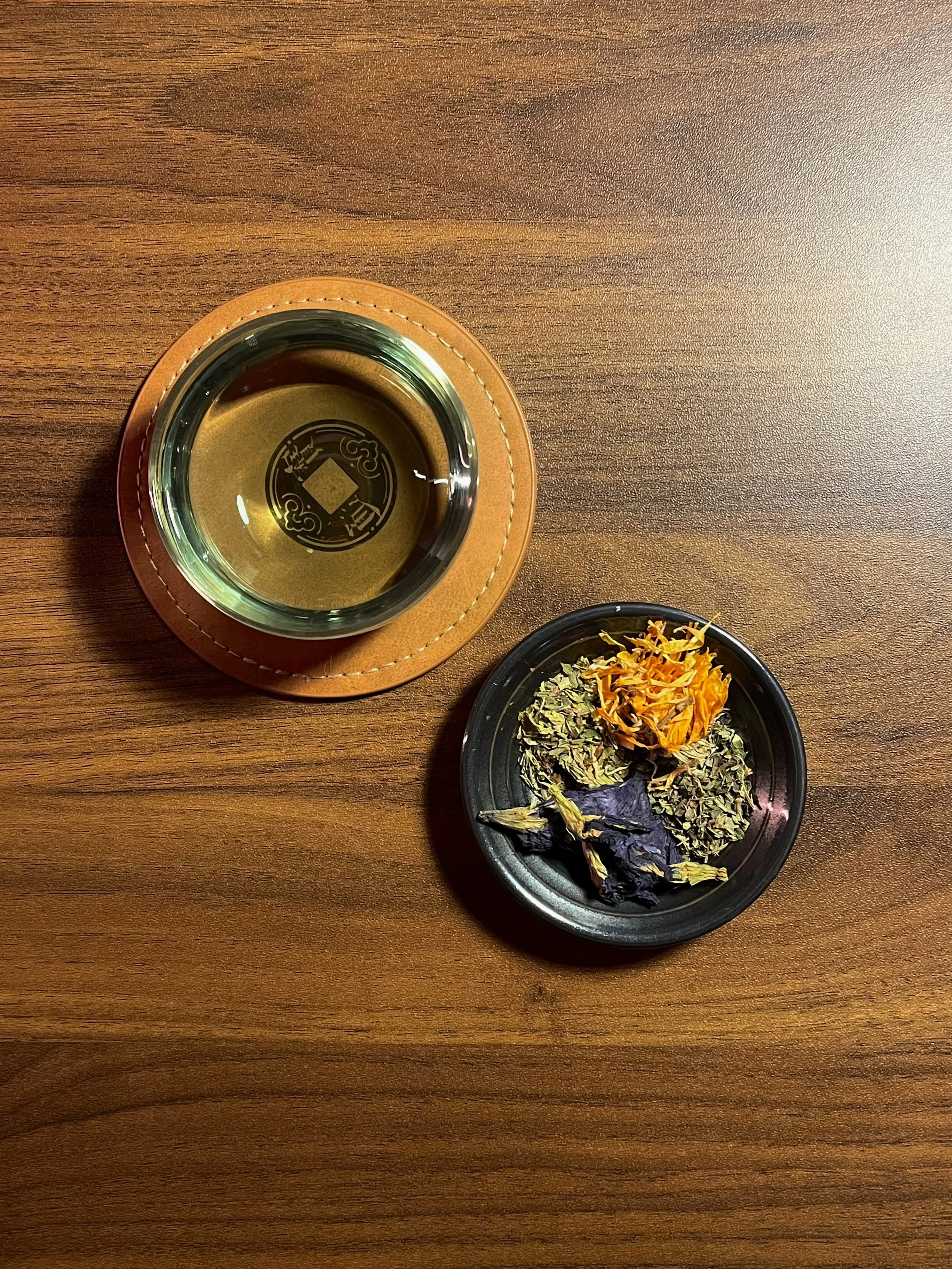 A wooden table with a glass of tea on a brown leather coaster and a black dish with dried flowers and herbs.