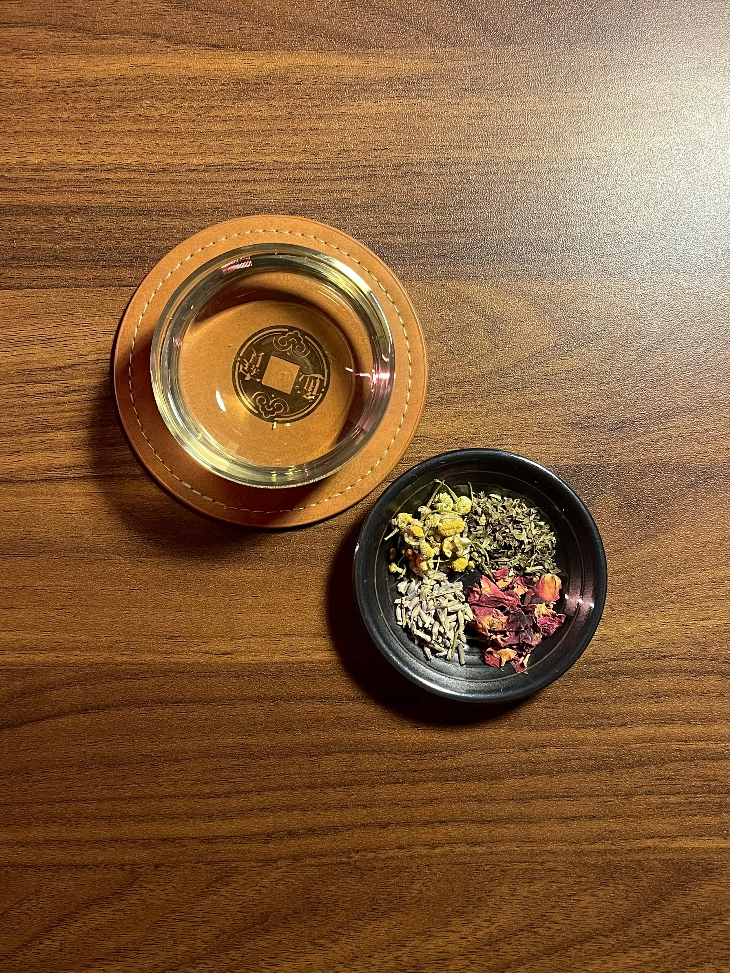 A wooden table with a glass of tea in a leather coaster, and a black dish filled with assorted dried flowers and herbs.