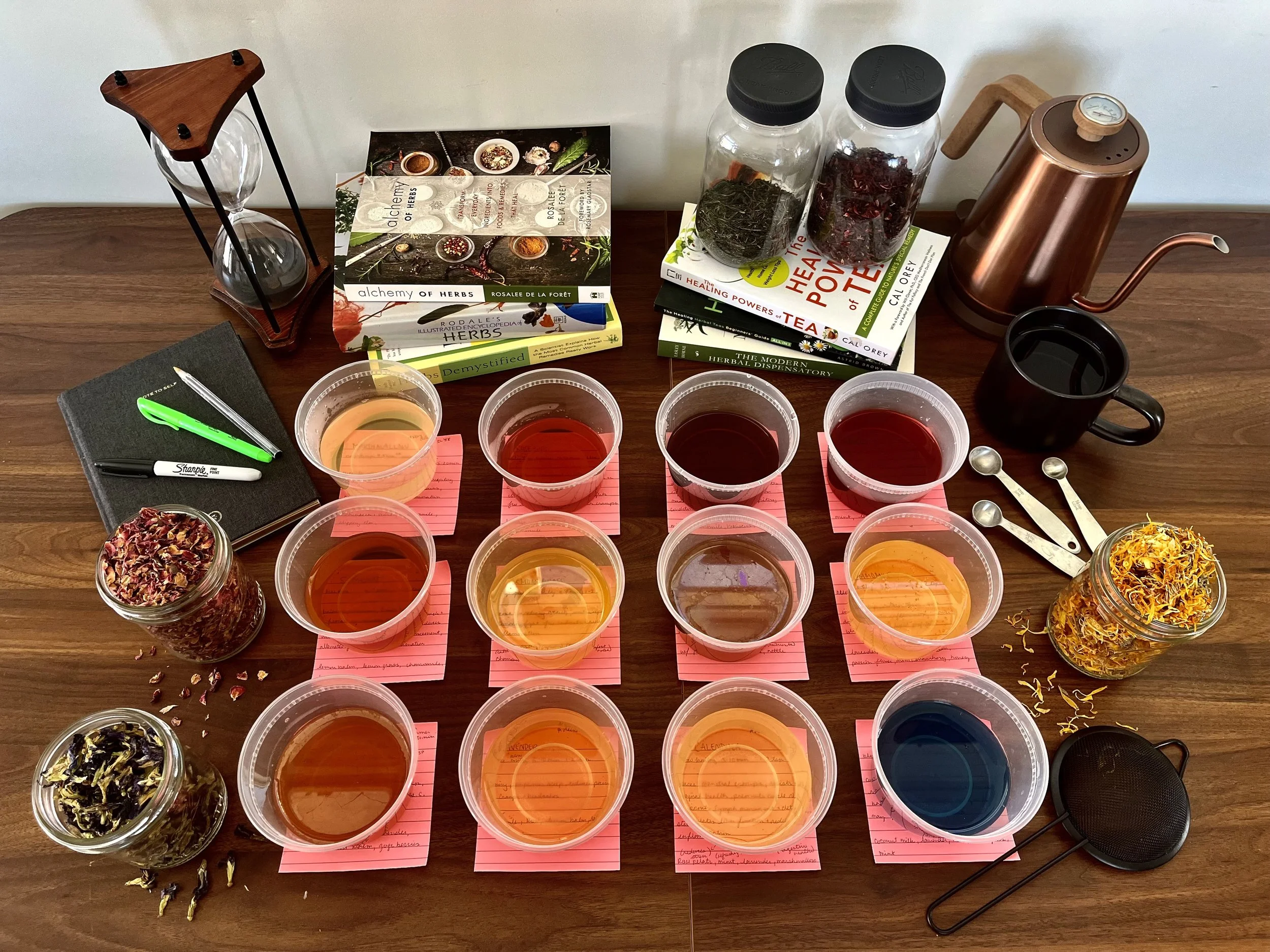 A wooden table with various jars of dried herbs, several small cups filled with different herbal infusions, a book titled "Alchemy of Herbs," a notepad with a green pen, and a copper tea kettle.