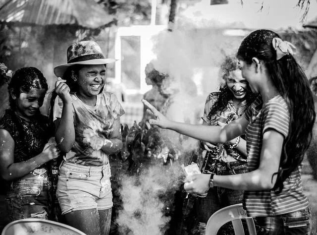 Four young girls are gathered around a small fire, smiling and laughing together outdoors, with one girl pointing at the fire and another holding an object.