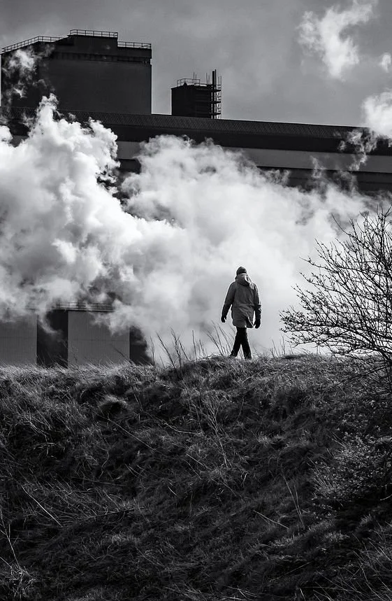 A person walking on a grassy hill near a large industrial building with smoke or steam billowing from it, in black and white.