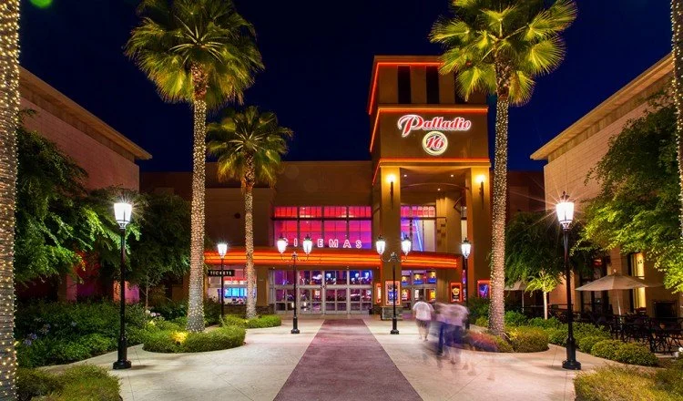 Nighttime view of an outdoor shopping center with palm trees, lit lamp posts, and a Palladio 16 Cinemas sign above its entrance.