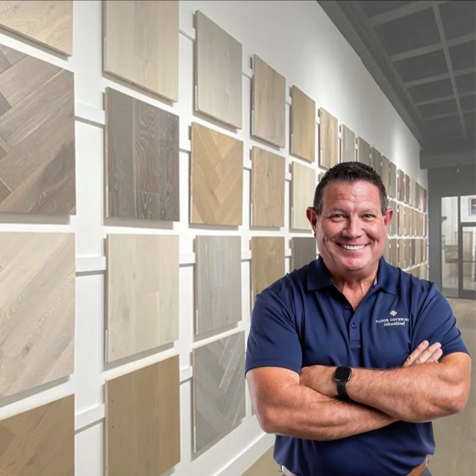 A man smiling with arms crossed, wearing a blue polo shirt, stands in front of a wall display of various wood flooring samples in an interior showroom.