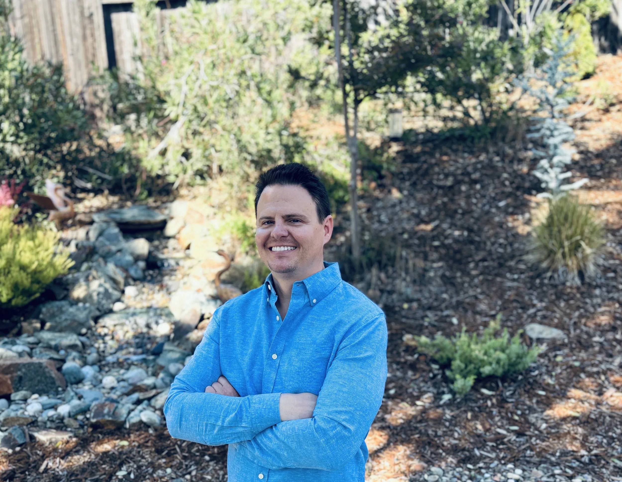 Smiling man in a blue button-up shirt with arms crossed, standing outdoors in a garden with various plants, rocks, and a small waterfall.