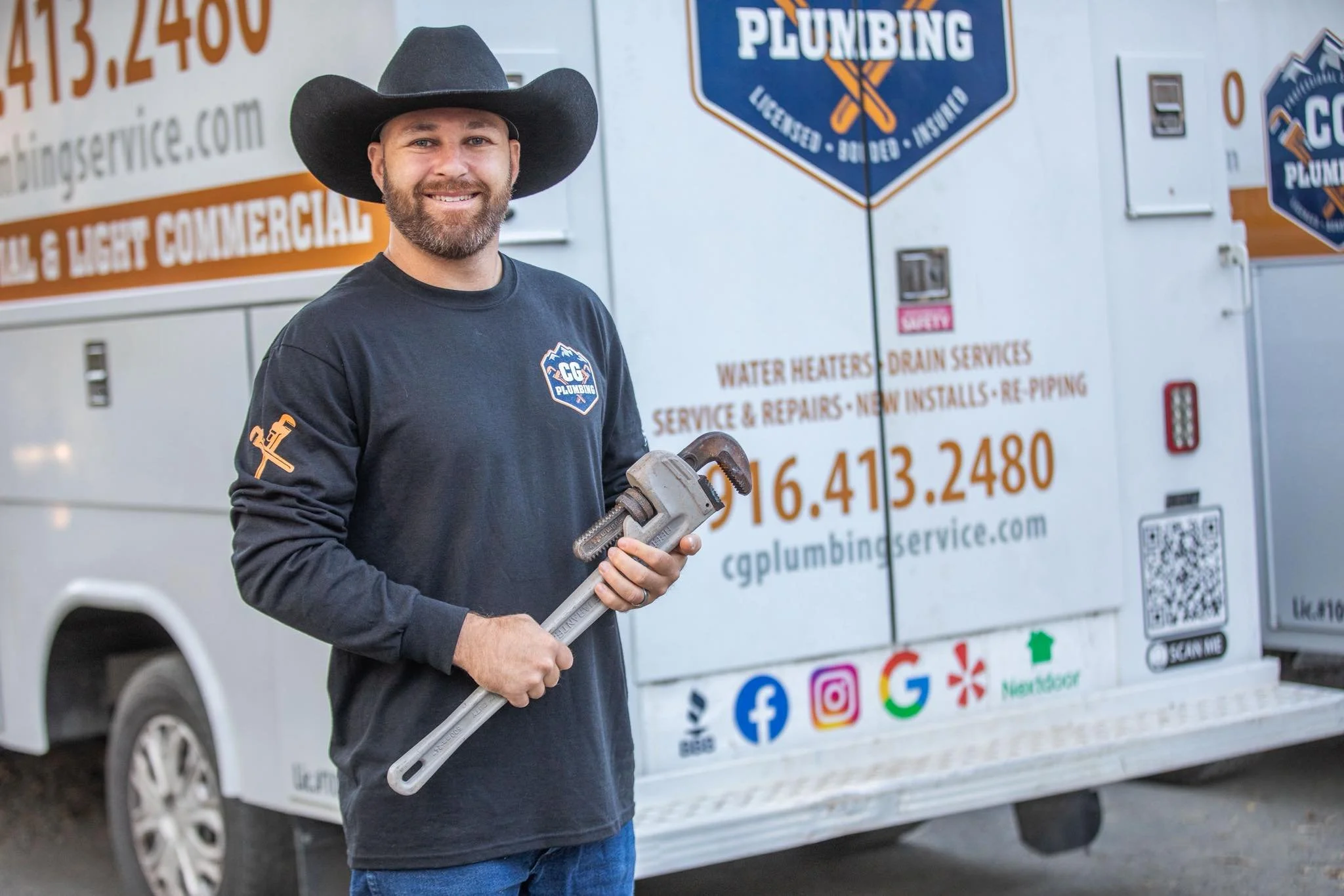 A man in a black long-sleeve shirt and cowboy hat holding a pipe wrench, standing in front of a plumbing service truck with company branding and contact information.