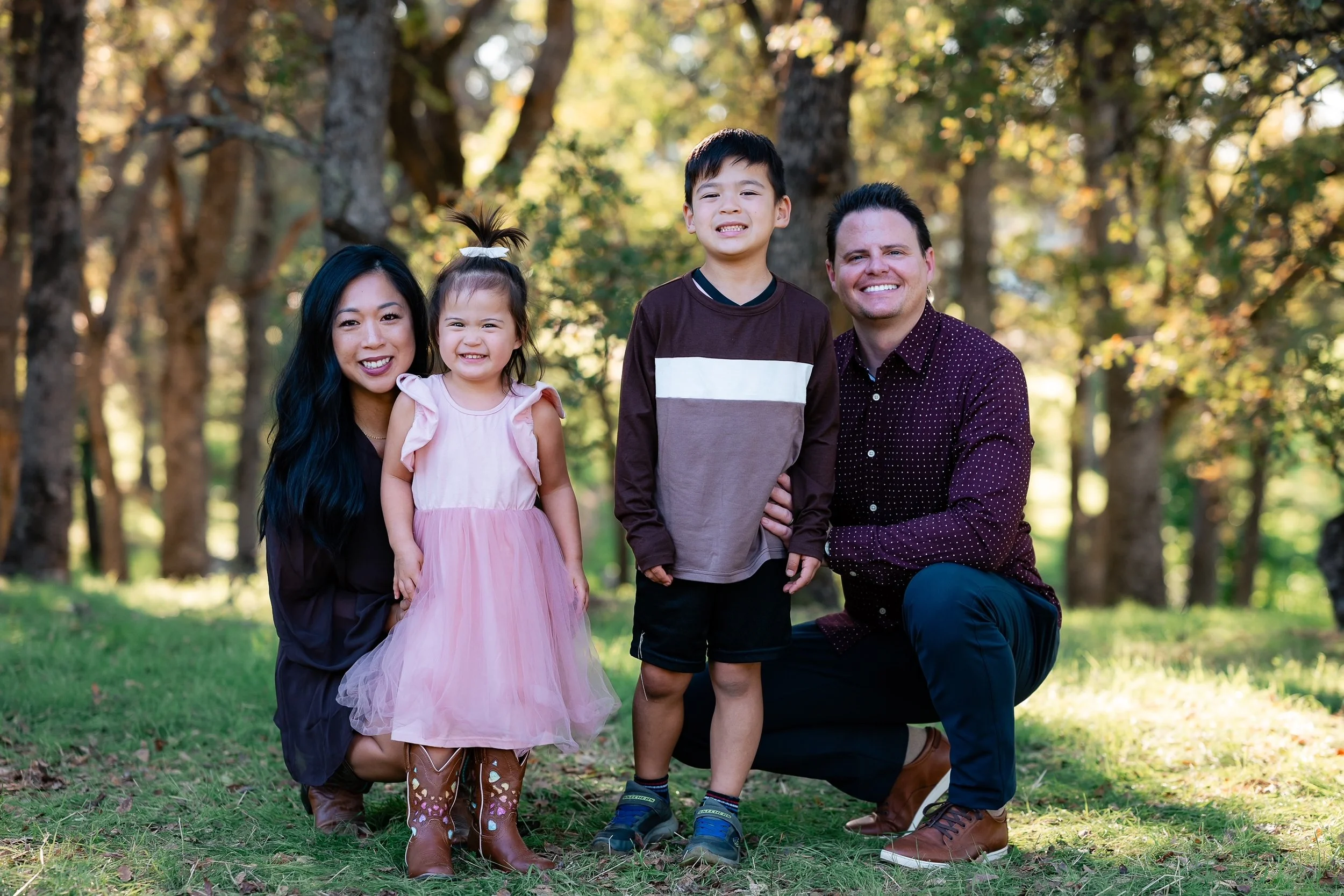 A family of four smiling in a wooded park, with a woman, a young girl in a pink dress, a boy in a brown shirt, and a man, all standing on grass during daytime.