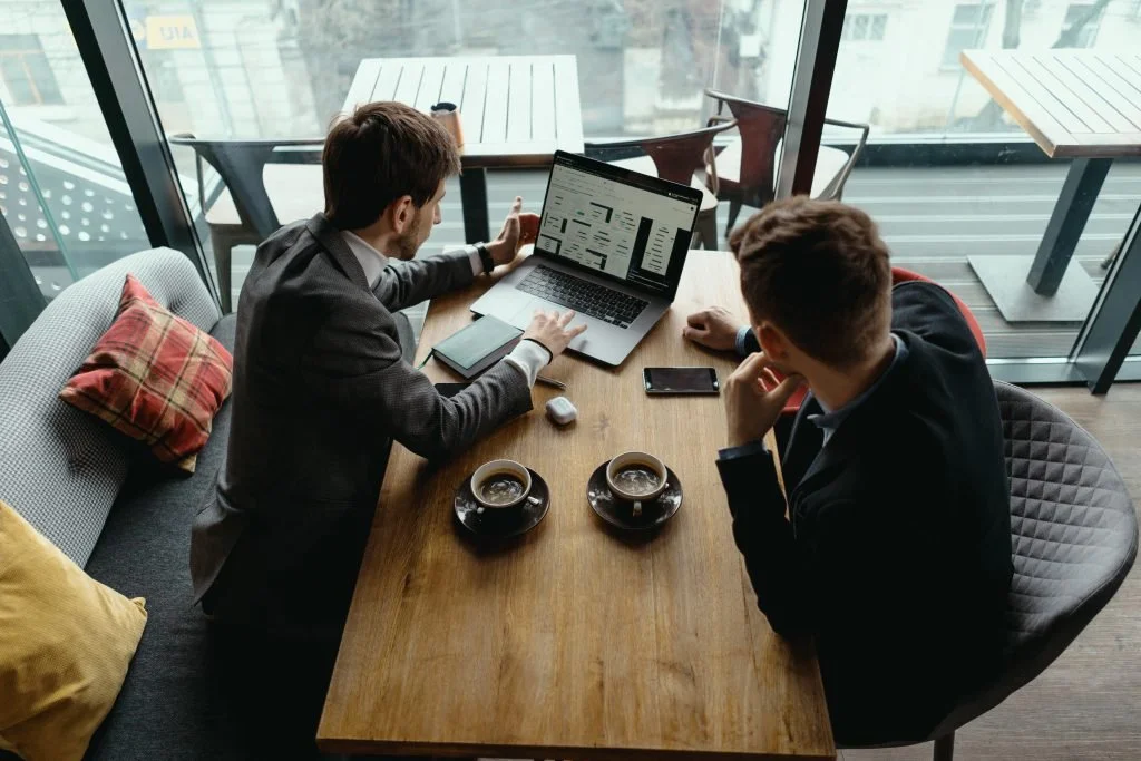 Two men sit at a wooden table in a coffee shop, engaging in a discussion with a laptop and smartphones on the table. There are two cups of coffee, a notebook, and a small device on the table. The coffee shop has large glass windows, revealing outdoor tables and a cityscape outside.