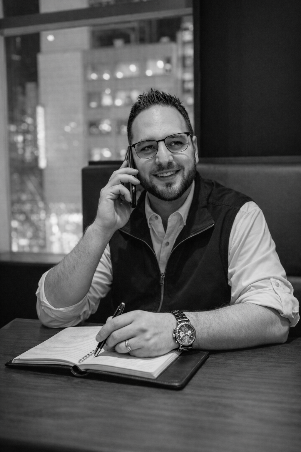 A man with glasses, a beard, and a watch, smiling while talking on a mobile phone, seated at a table with an open notebook and a pen in an urban setting.