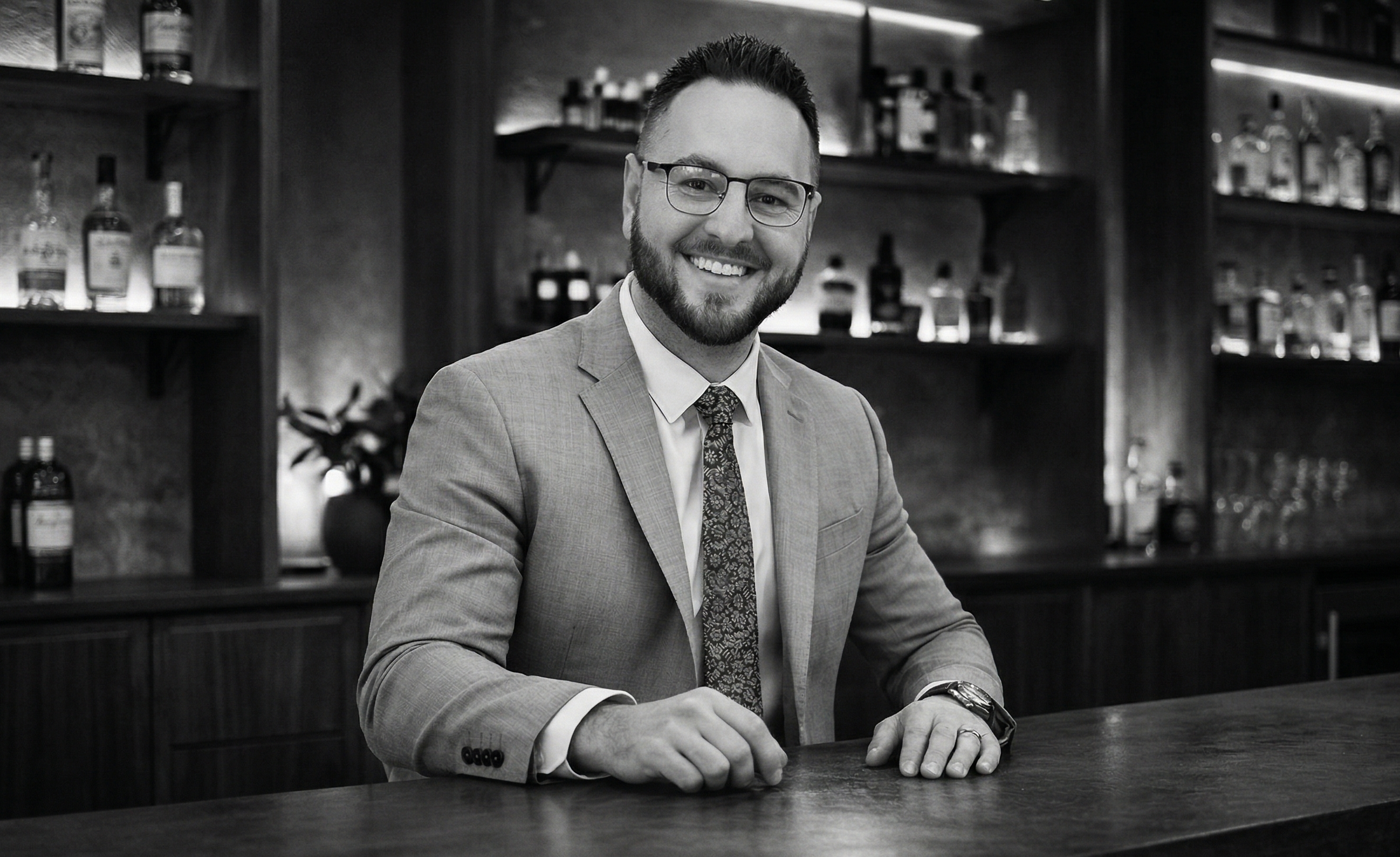 A smiling man in a suit and tie sitting at a bar, with shelves of liquor bottles in the background.