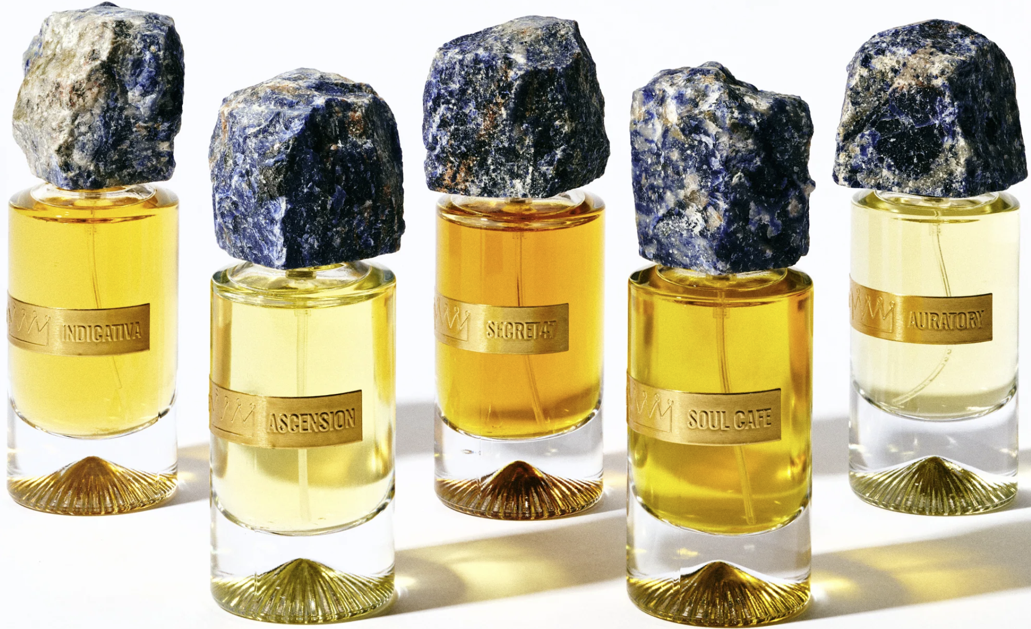 Five small glass bottles of perfume with blue stones on top, arranged in a row on a white background. Each bottle has a gold label with words indicating different perfume names.
