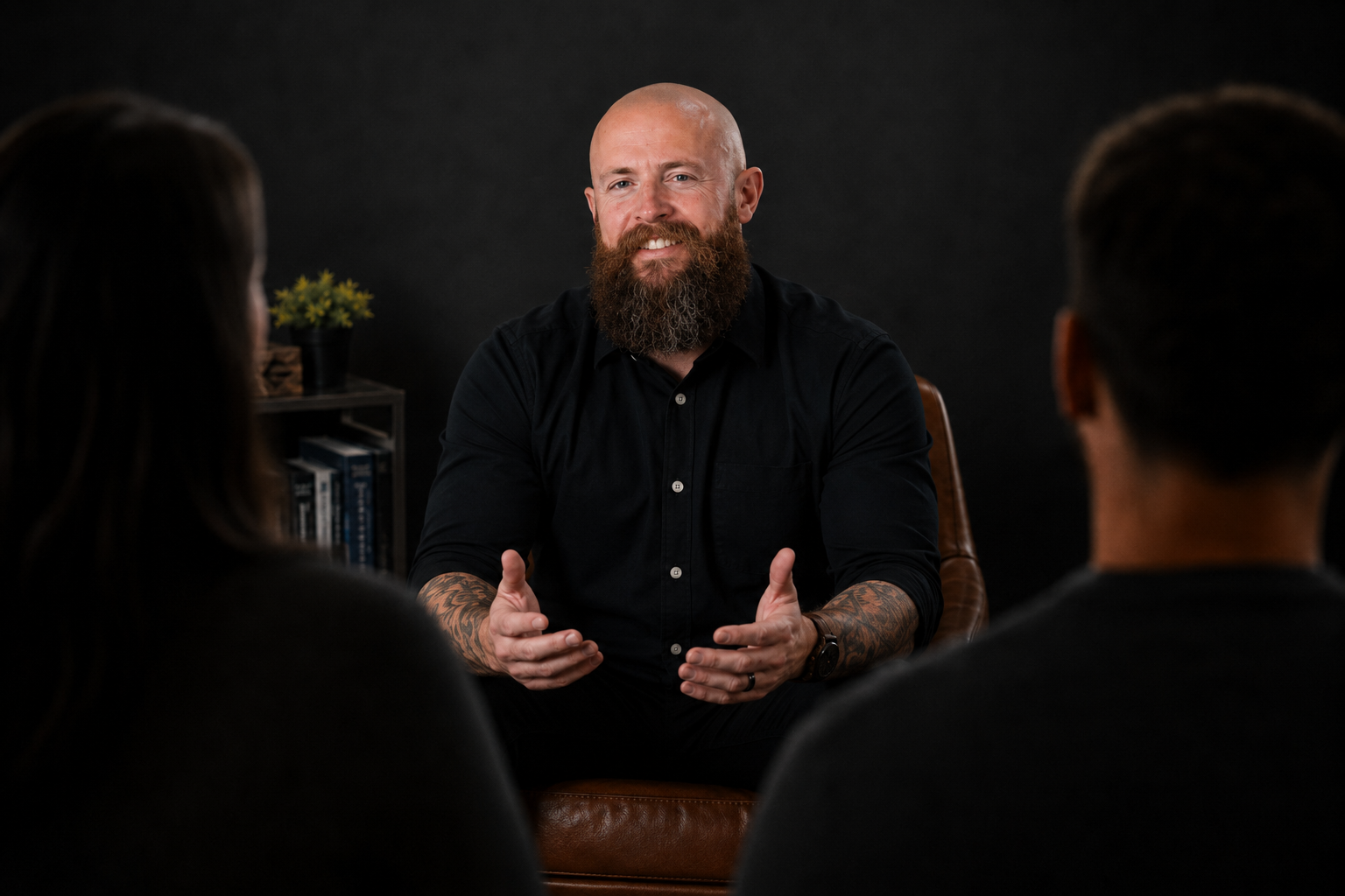 A man with a beard and tattoos on his arms, wearing a black shirt, is speaking to two people with their backs to the camera in a dark, professional setting.