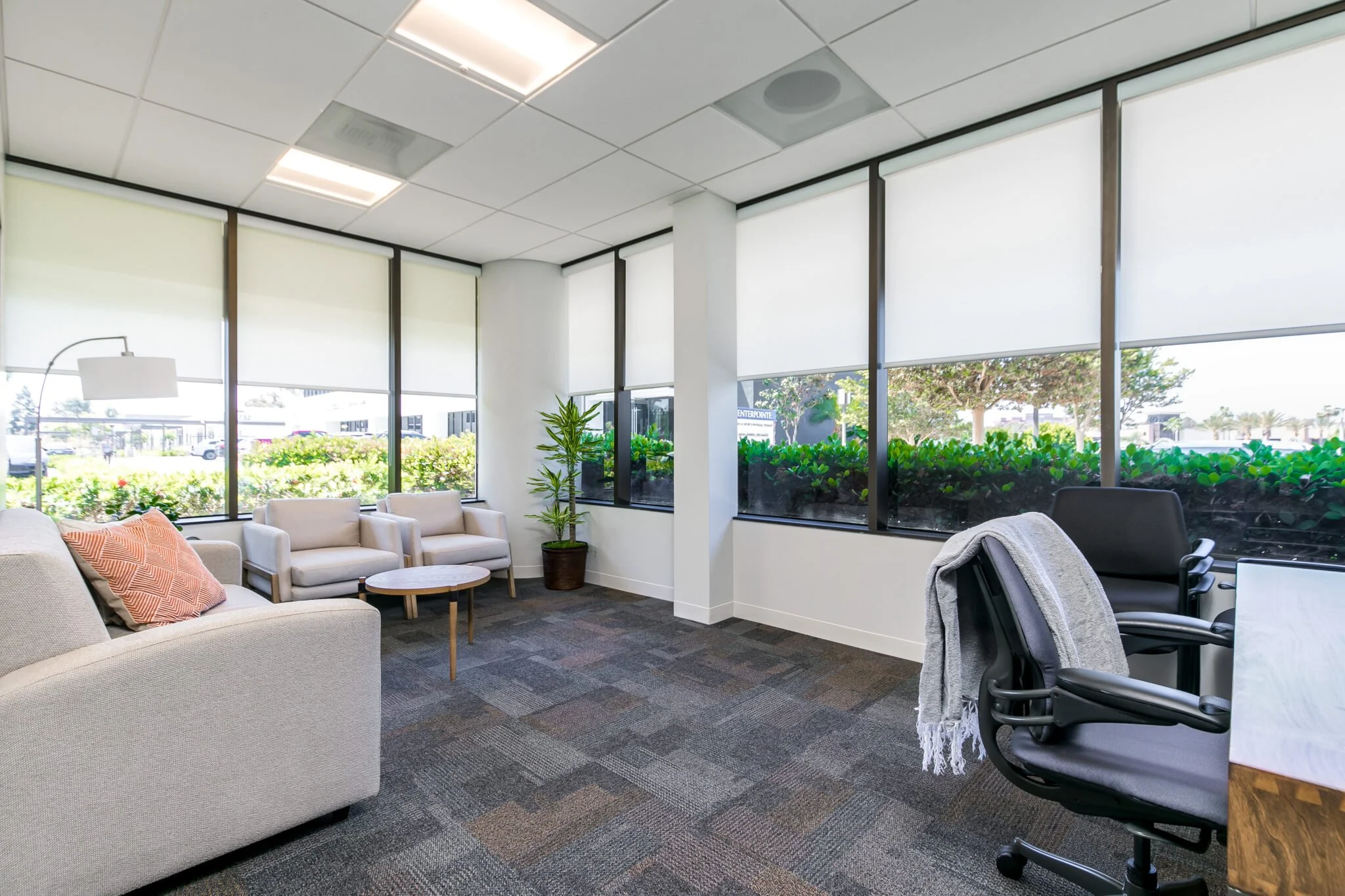 An office lounge area with beige sofas, a small round wooden table, plants, large windows with blinds, and office chairs, with view of parking lot and trees outside.