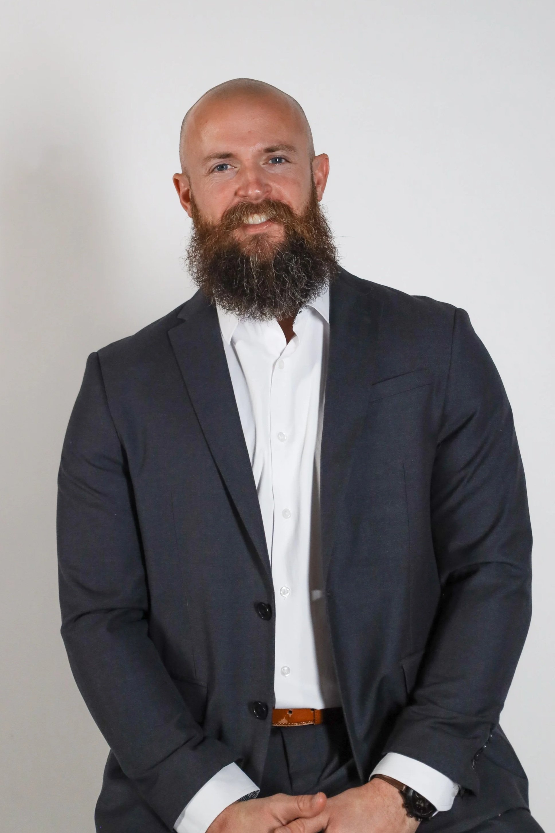 A man with a beard and a shaved head, dressed in a dark suit and white shirt, sitting against a plain white background.