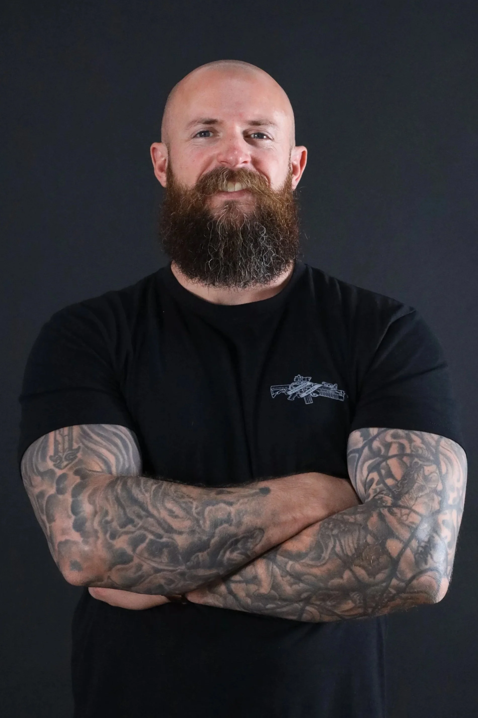 A bald man with a full beard and tattoos on his arms, crossing his arms in front of a dark background, wearing a black shirt with a graphic of a gun.