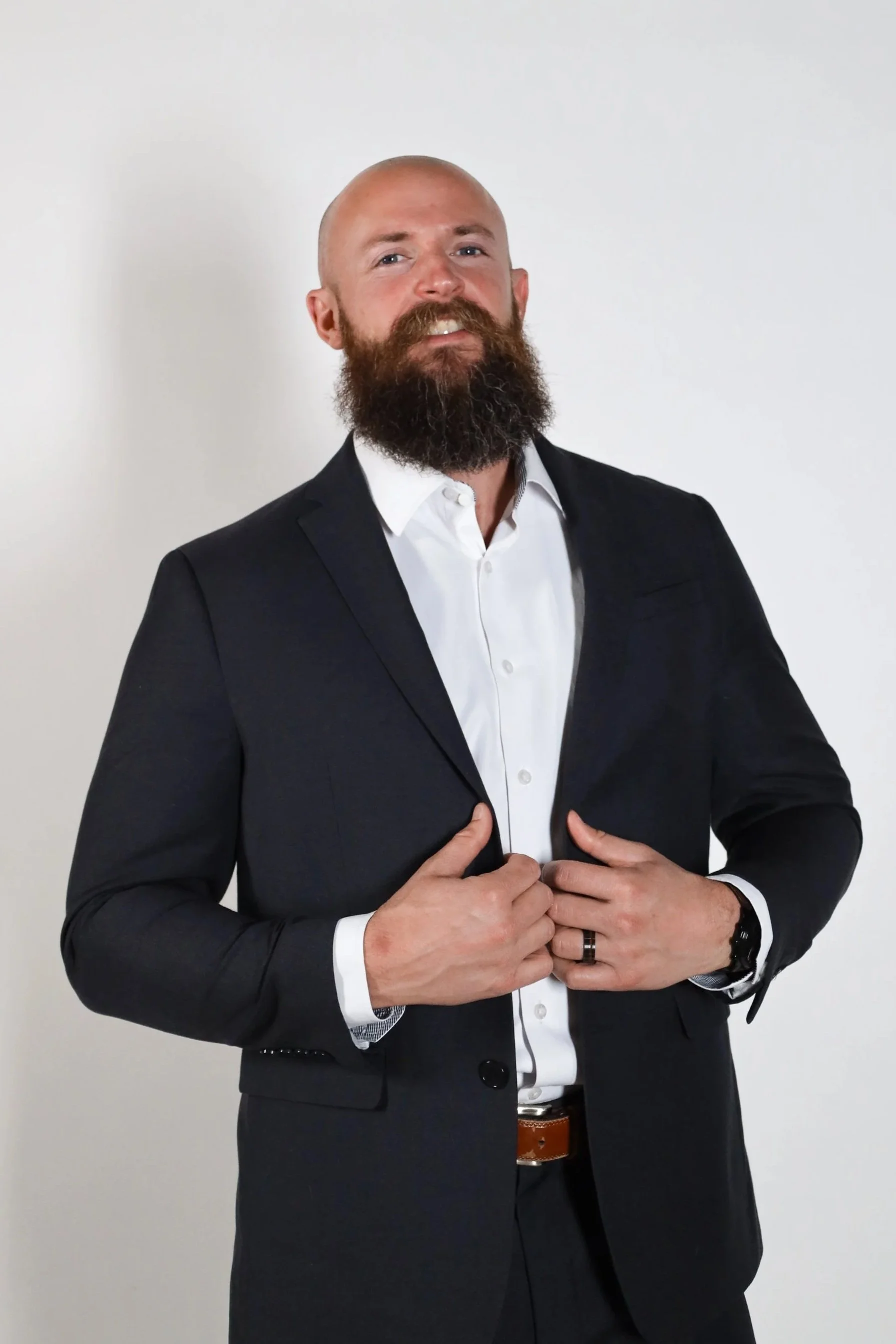 A man in a black suit with a white dress shirt, standing against a plain white background. He has a bald head, a full beard, and is adjusting his suit jacket with both hands.