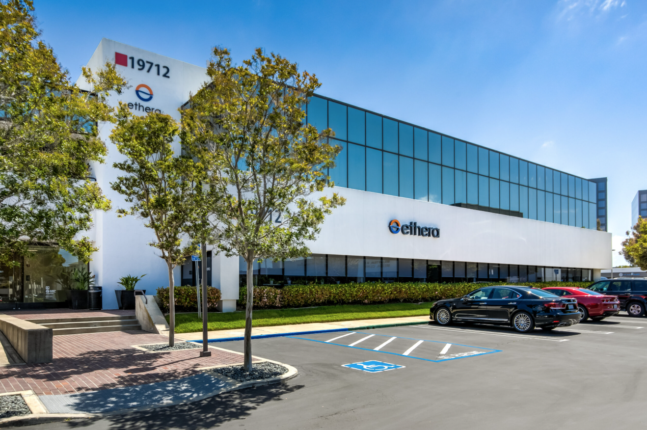 Modern office building with glass windows and a white facade, featuring the Ethera logo. There are parking spaces in the foreground, including a reserved handicapped spot, and a few parked cars. Green trees line the sidewalk under a clear blue sky.