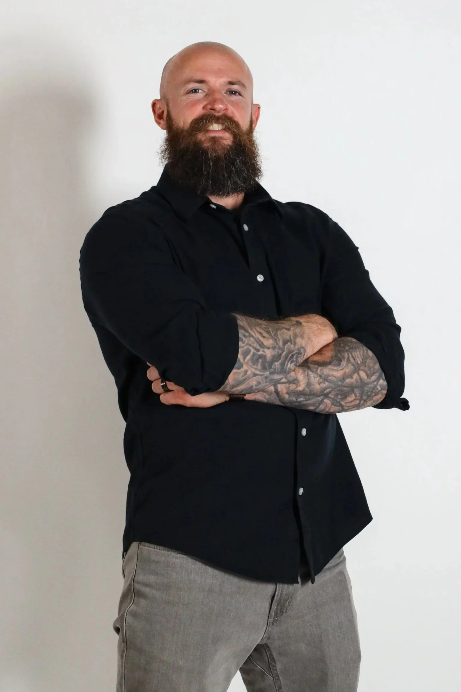 A man with a beard and tattooed arms wearing a black button-up shirt and gray pants, standing with arms crossed against a plain white wall.