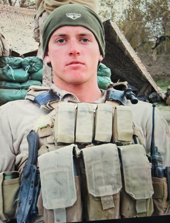 A soldier wearing a green beanie, tactical vest with pouches, and a beige uniform, standing outdoors with military gear in the background.