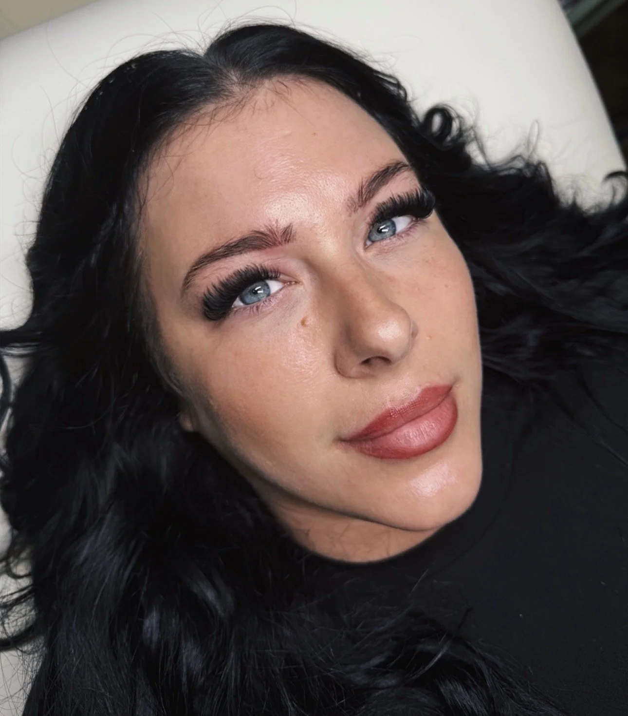 Close-up of a woman with long black hair, blue eyes, and makeup, lying down and looking at the camera.