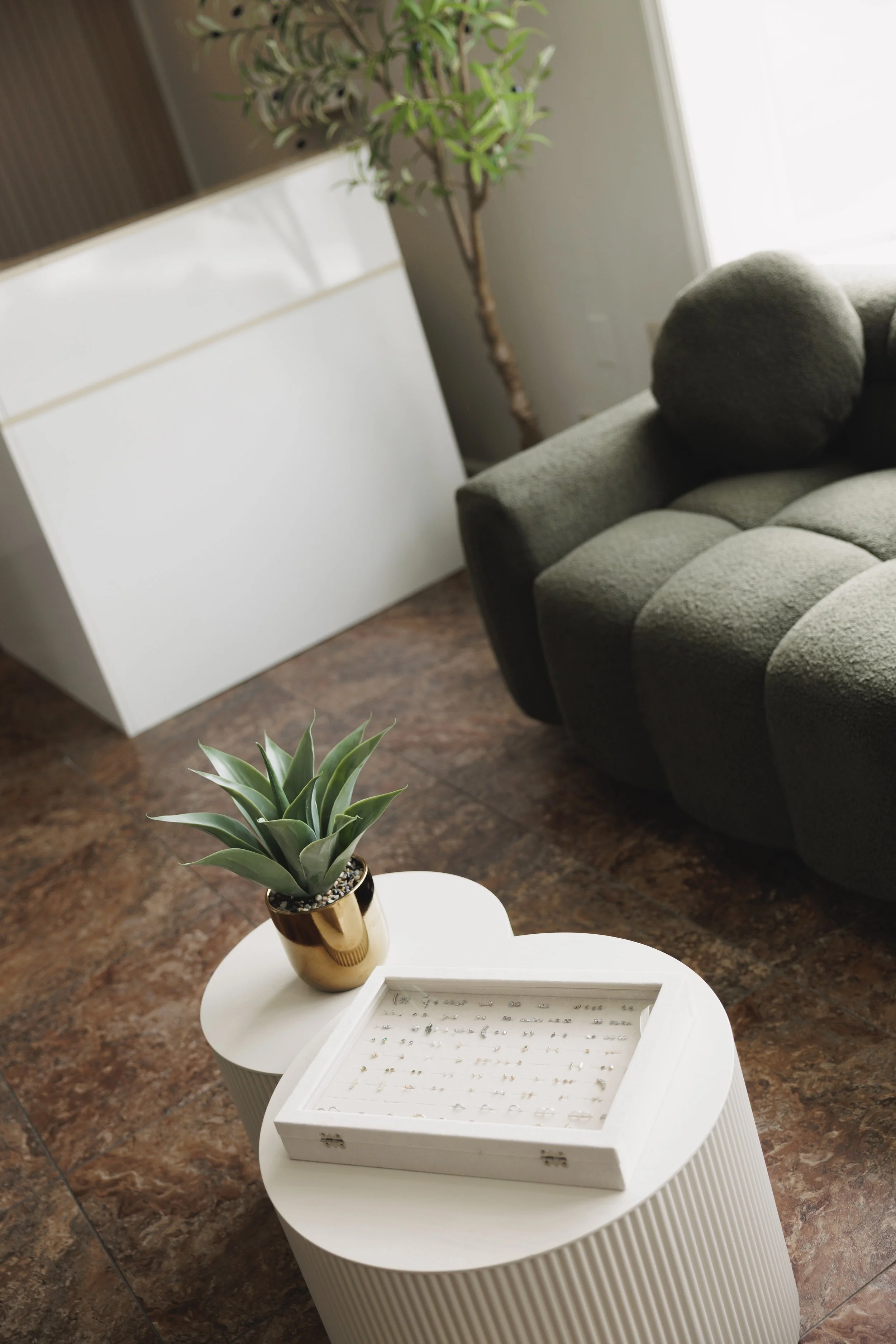 A living room with a dark green upholstered sofa, a small round white side table with a plant in a shiny gold pot, and a white jewelry box on the table. A tall potted tree stands in the background near a window, and a white cabinet is next to it. The floor is brownish tile.