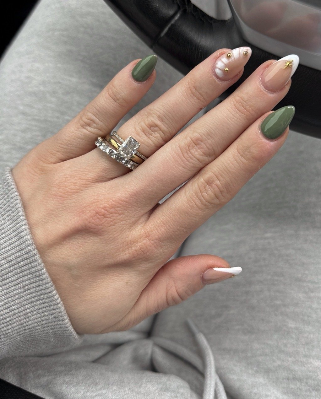 A hand with manicured nails wearing a ring with a large rectangular diamond and gold accents. The nails are painted in dark green, white with pearl embellishments, and nude with a gold star, set against a gray fabric background.