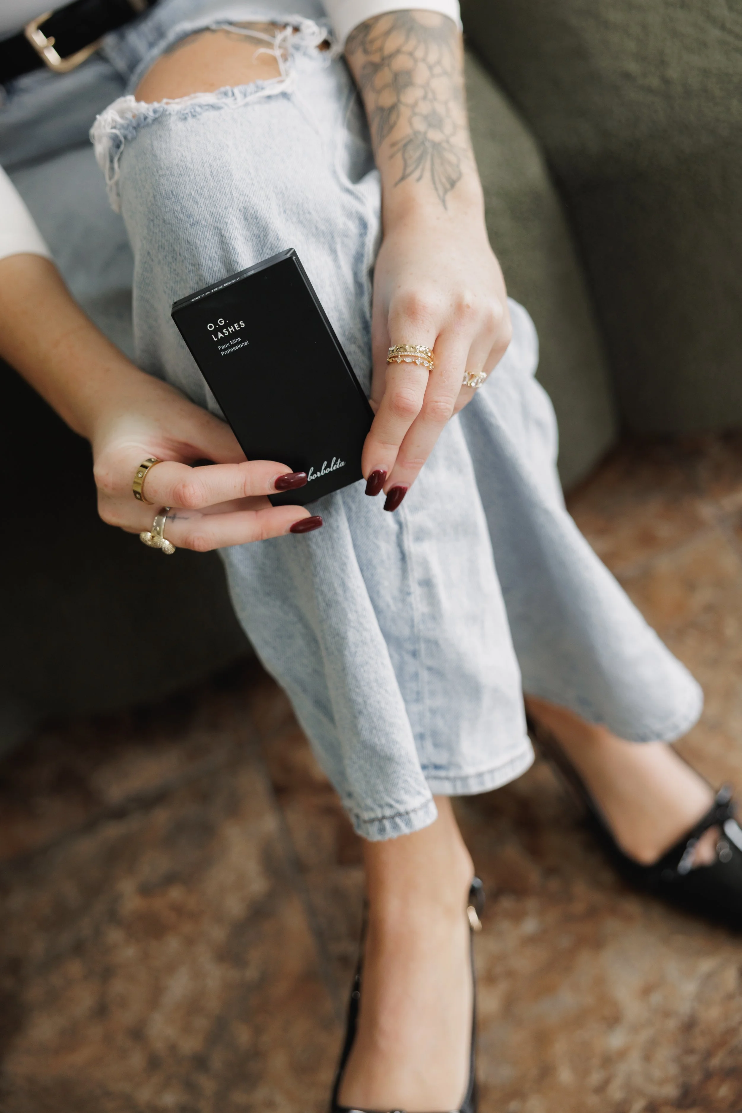 Person sitting on a couch holding a black box of false eyelashes, wearing ripped light-wash jeans, black shoes, and various rings, with a tattoo on their arm.