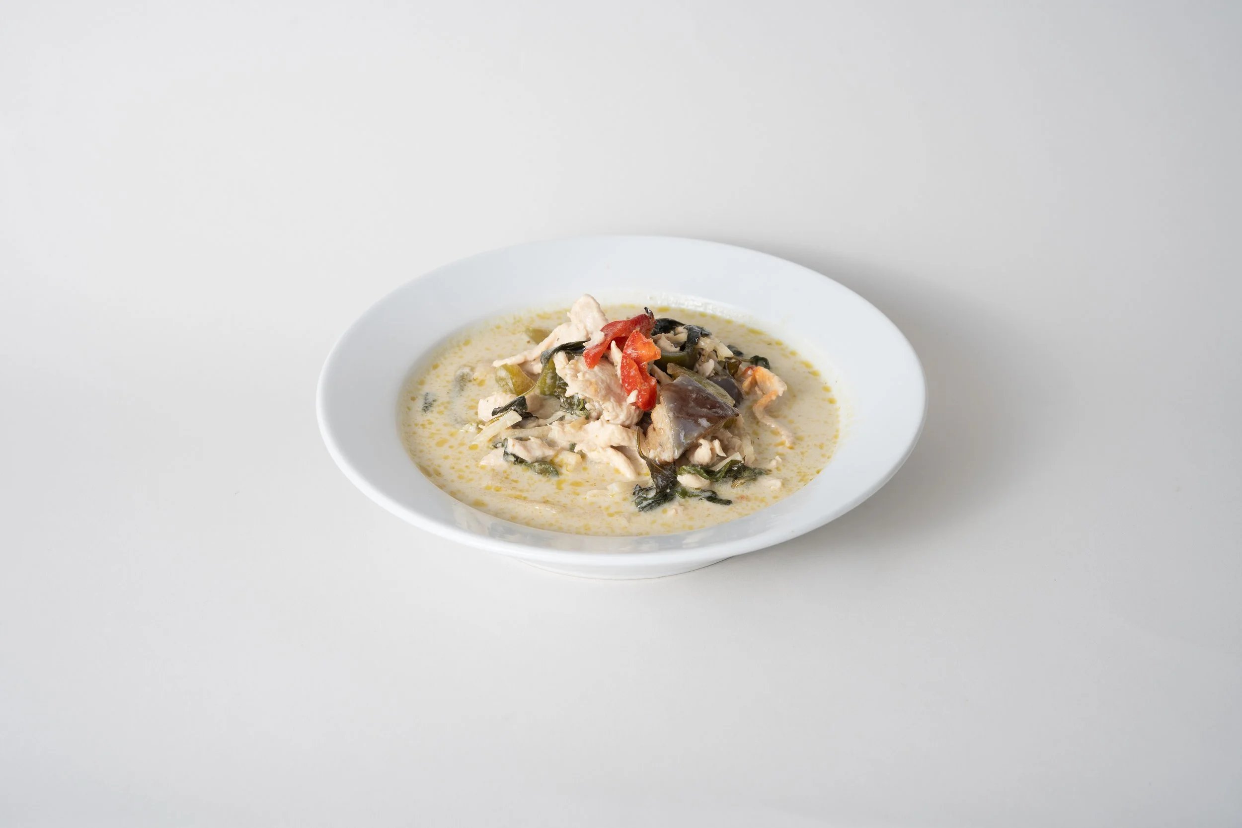 A white bowl of chicken and vegetable curry with green herbs and red peppers on a white surface.