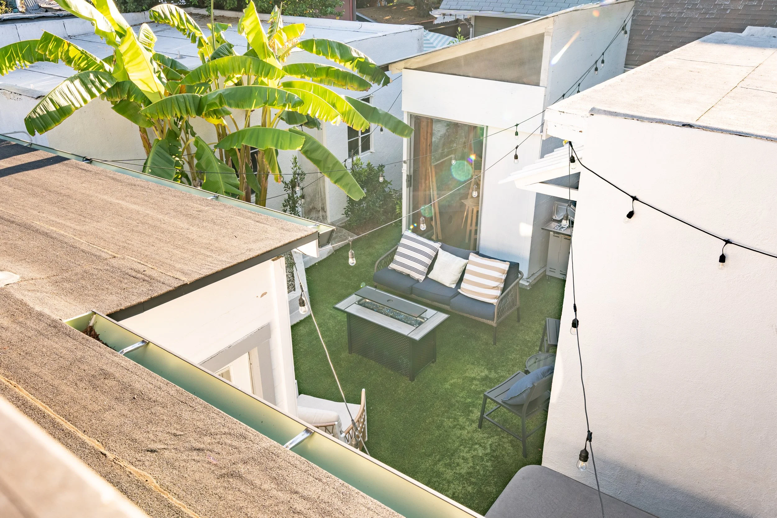 Small backyard patio with artificial grass, a black outdoor sofa with striped and solid pillows, a glass-top fire pit table, a gray side chair with pillows, string lights hung overhead, and a large banana plant in the corner.