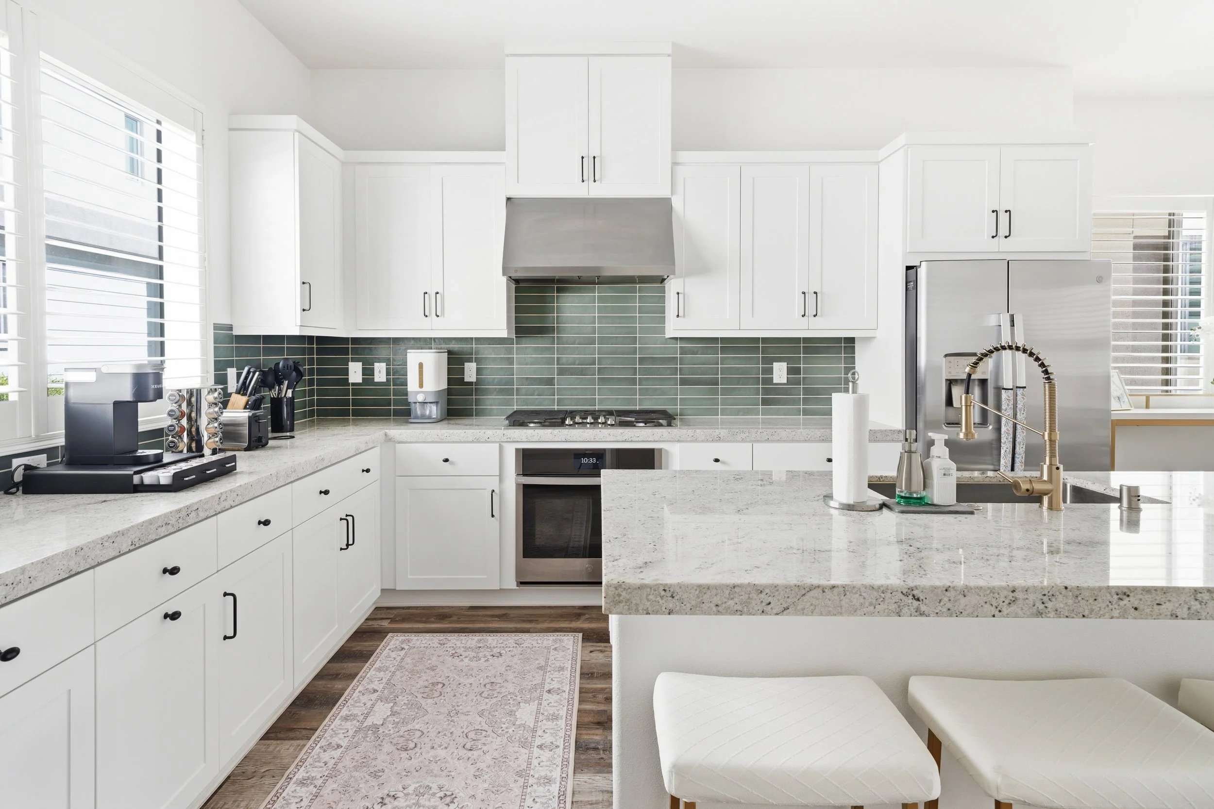 Modern kitchen with white cabinets, granite countertops, green backsplash tiles, stainless steel appliances, and a bar with white stools.