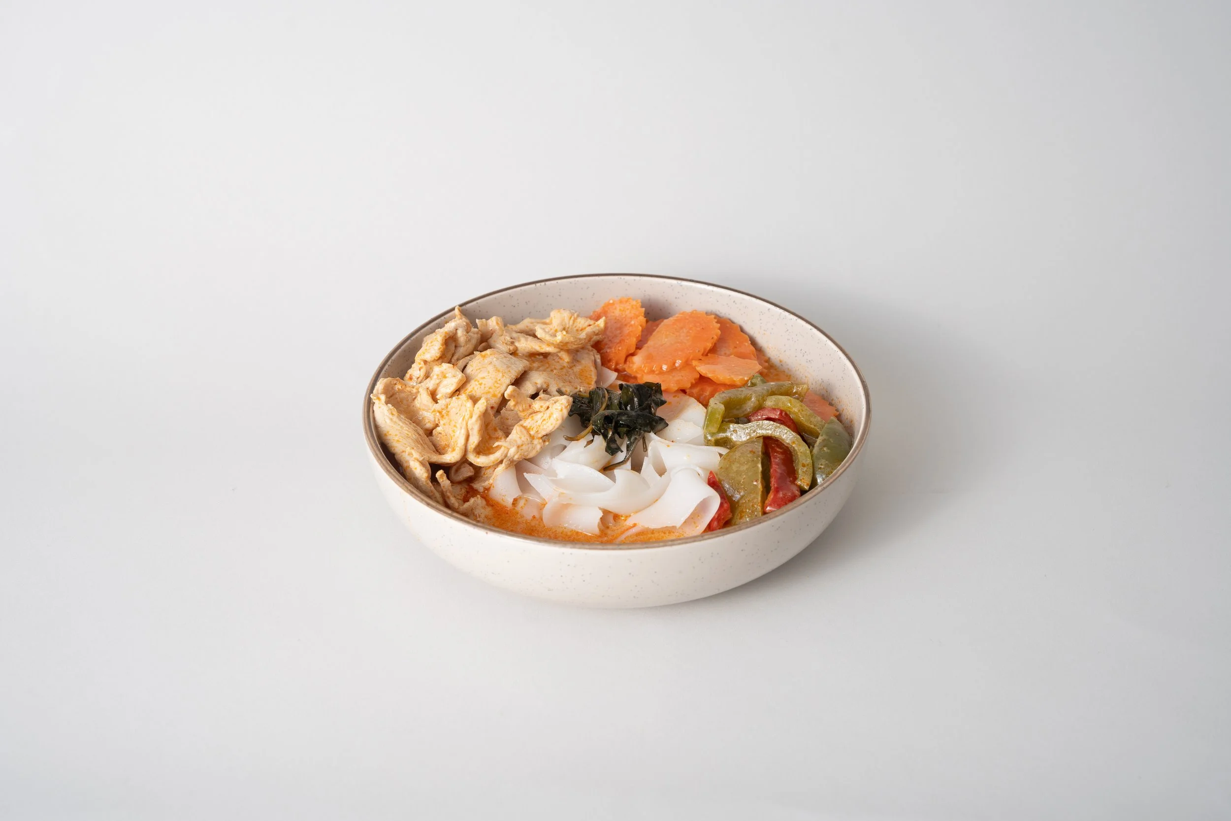 Bowl of Asian soup with sliced chicken, rice noodles, carrots, green and red bell peppers, and seaweed on a light background.