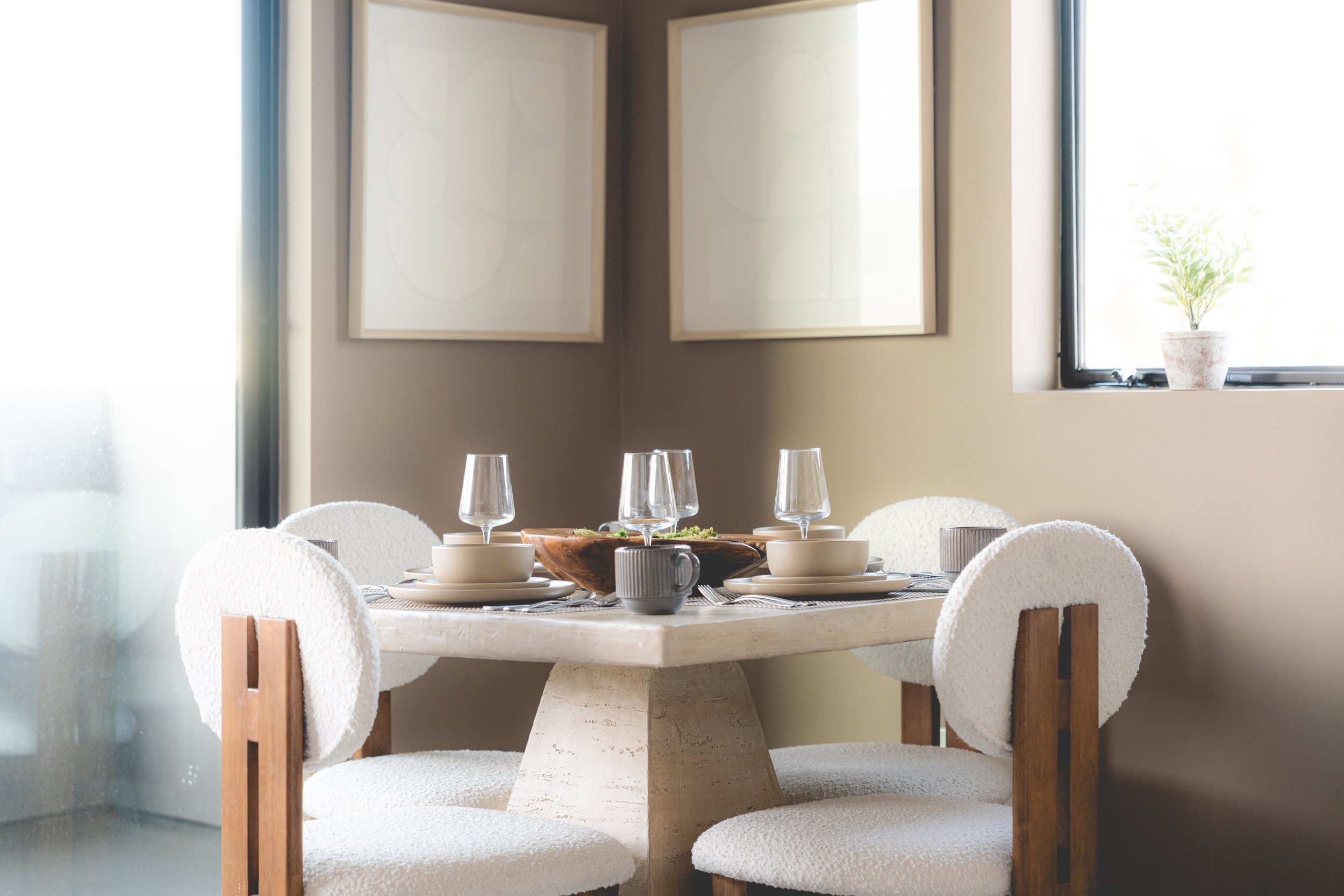Dining room with a round table set with plates, bowls, glasses, and a bowl of salad, four white cushioned chairs, a window with a potted plant, and two large abstract paintings on the wall.