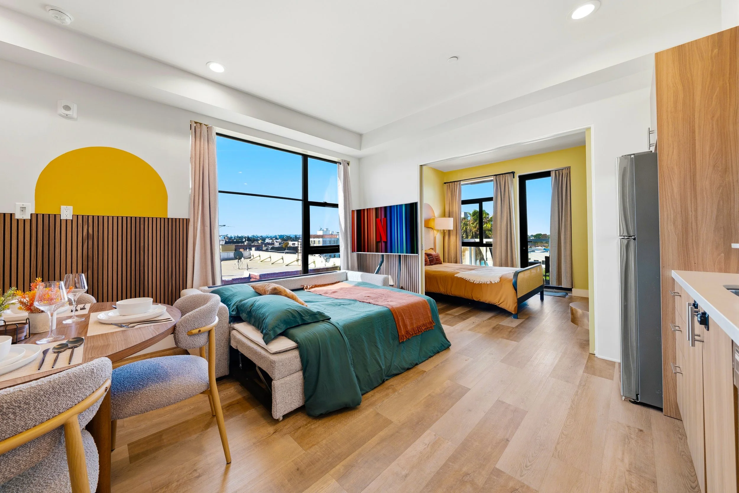 A modern open-plan apartment with two beds, a dining table, and a kitchen area. Large windows show an outdoor cityscape.