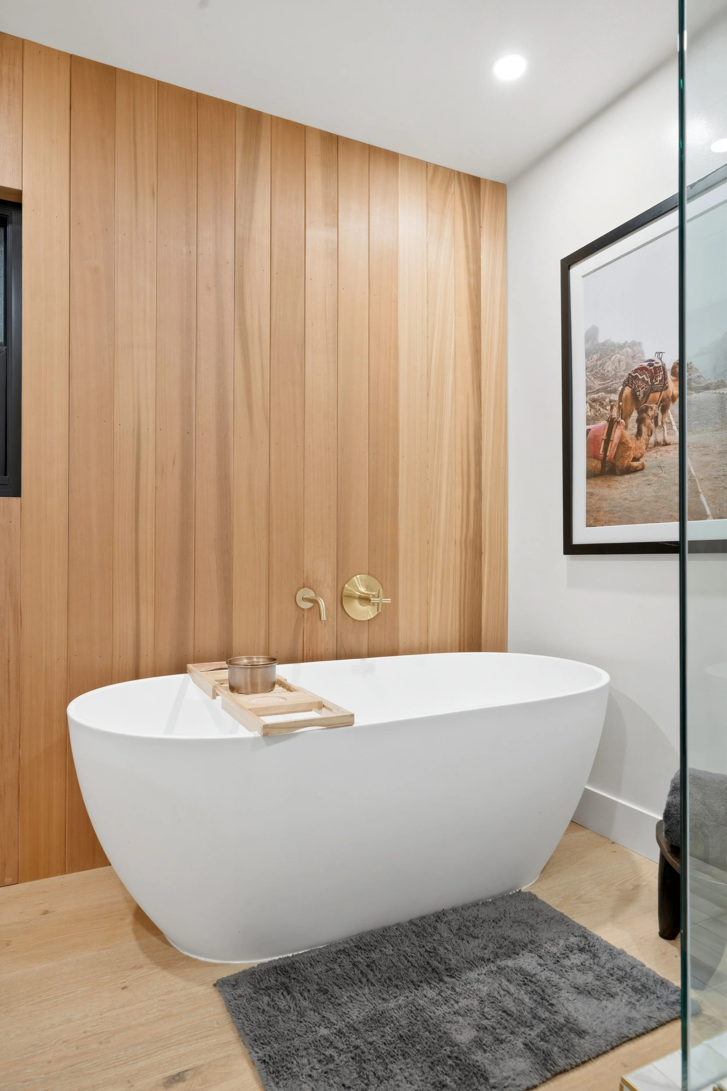 Modern bathroom with white freestanding bathtub, wooden accent wall behind it, framed artwork of a camel, gray rug, and minimalistic fixtures.