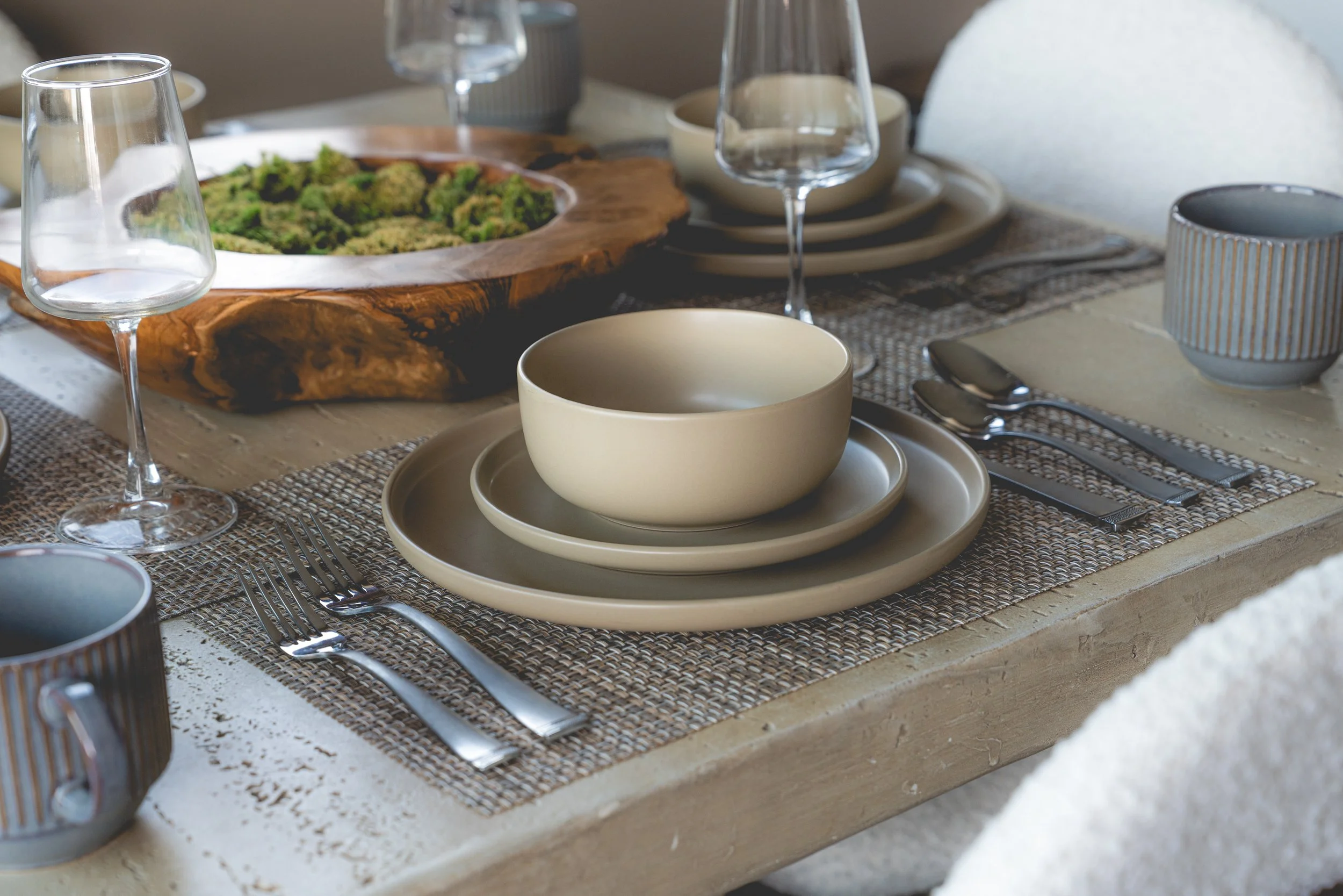 A neatly arranged dining table set for a meal with beige plates, bowls, forks, and knives on a woven placemat. There are several empty wine glasses and decorative bowls, with a large wooden serving platter holding a green salad in the background.