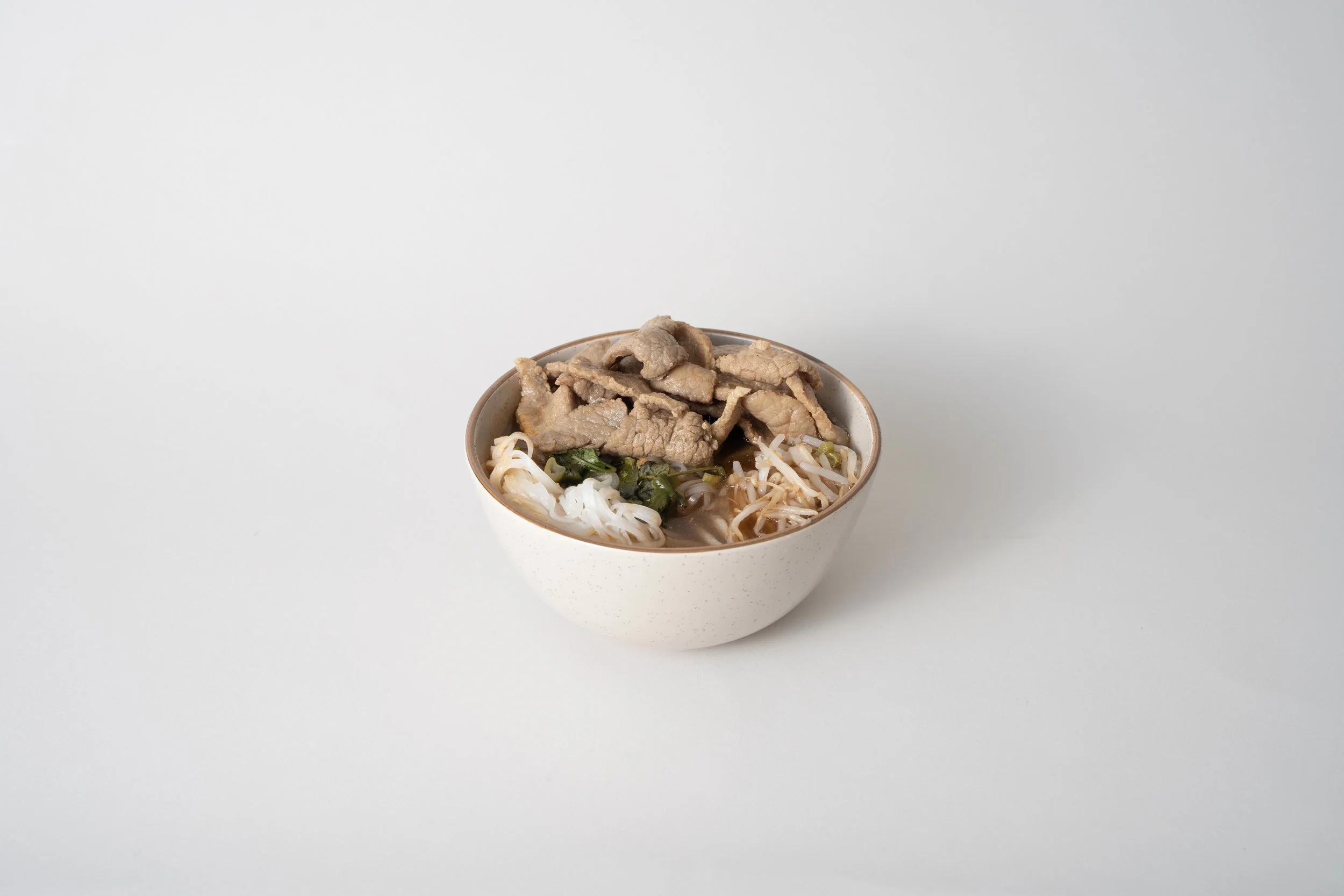 A bowl of Asian noodle soup with sliced meat, bean sprouts, and greens on a white background.