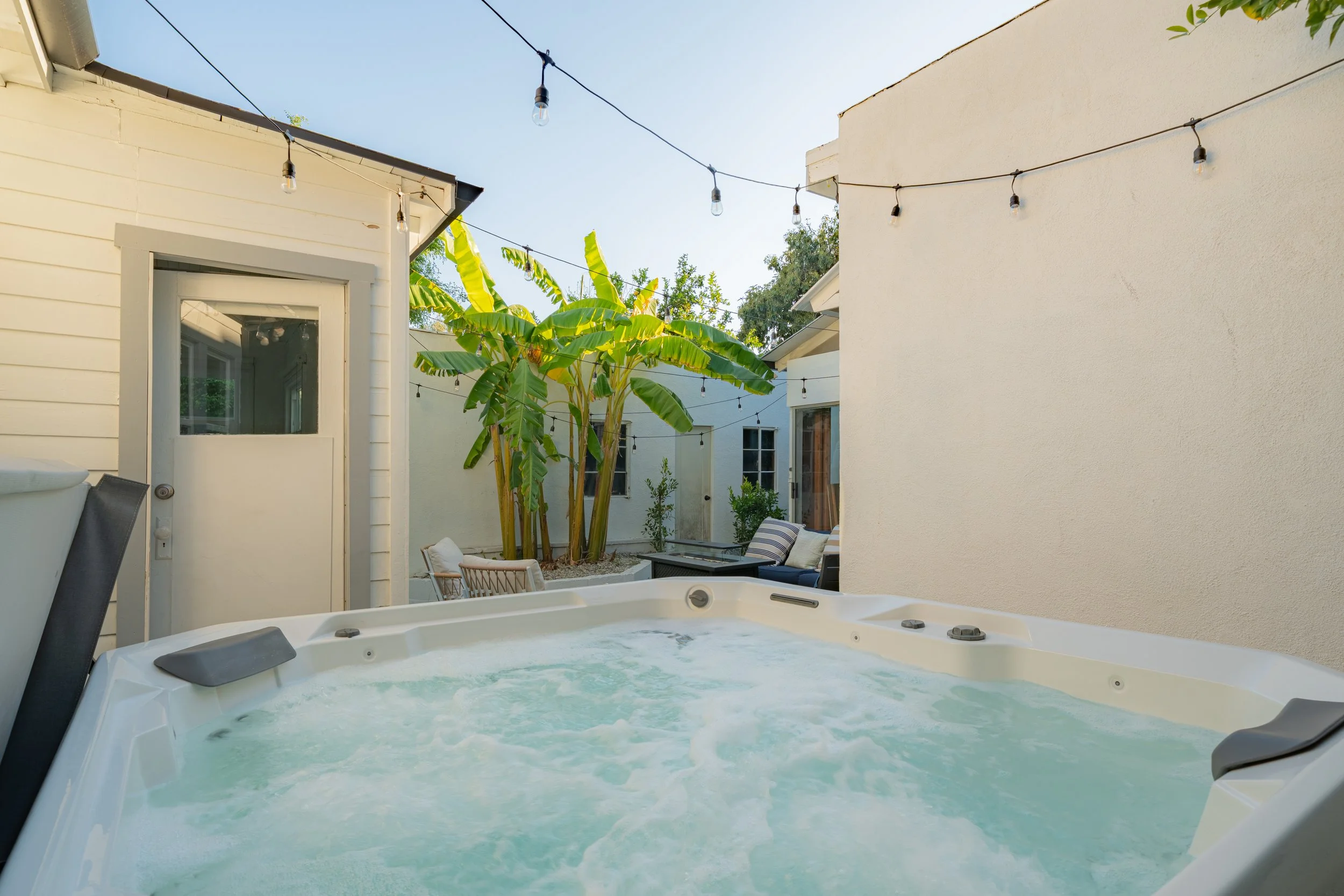 Outdoor backyard scene with a hot tub filled with bubbling water, string lights hanging overhead, banana trees, patio furniture, and a bright sky.