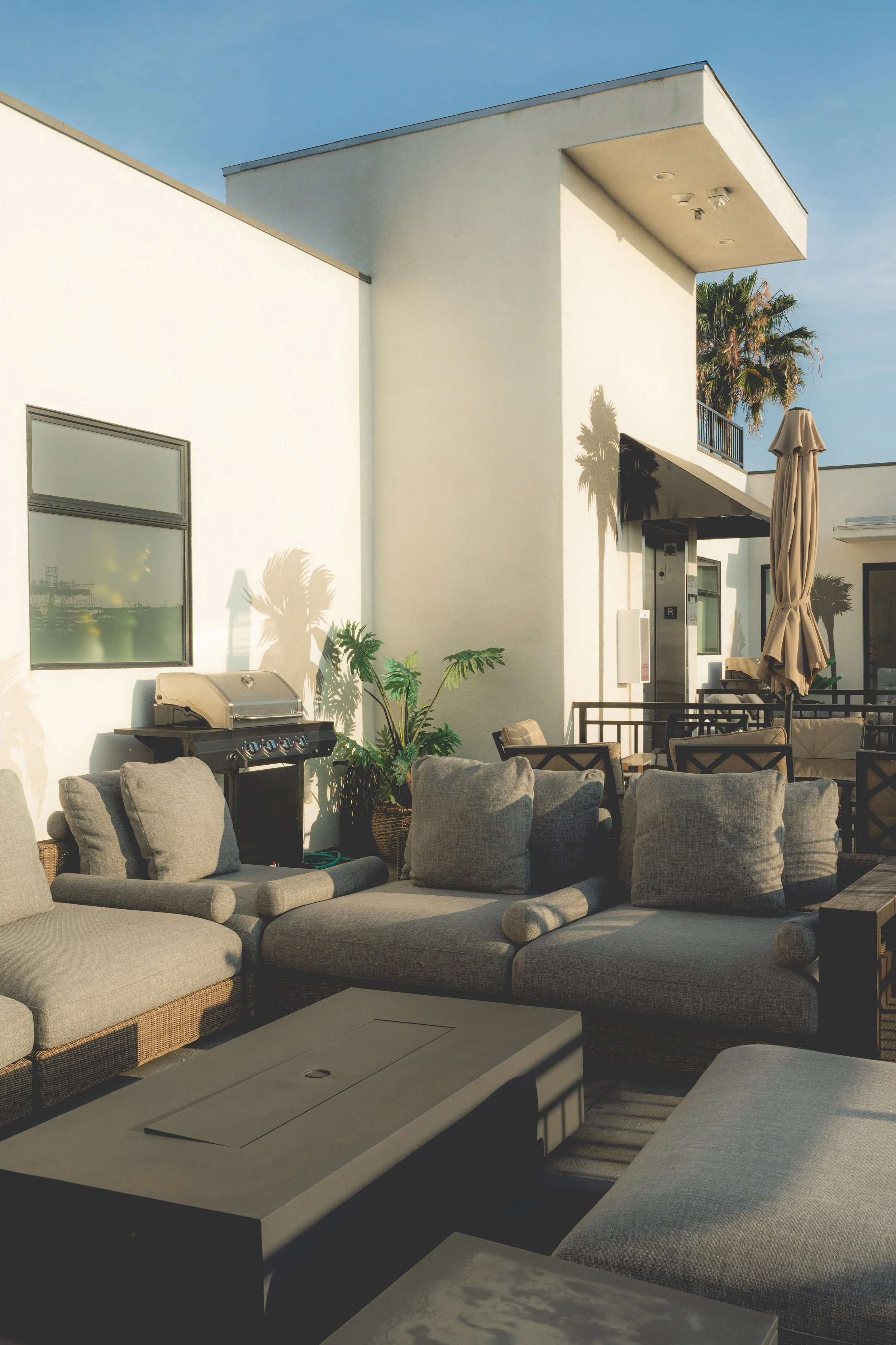 An outdoor patio area with beige patio furniture, a grill, potted plants, and a modern white building with a flat roof and balcony, under a clear blue sky.
