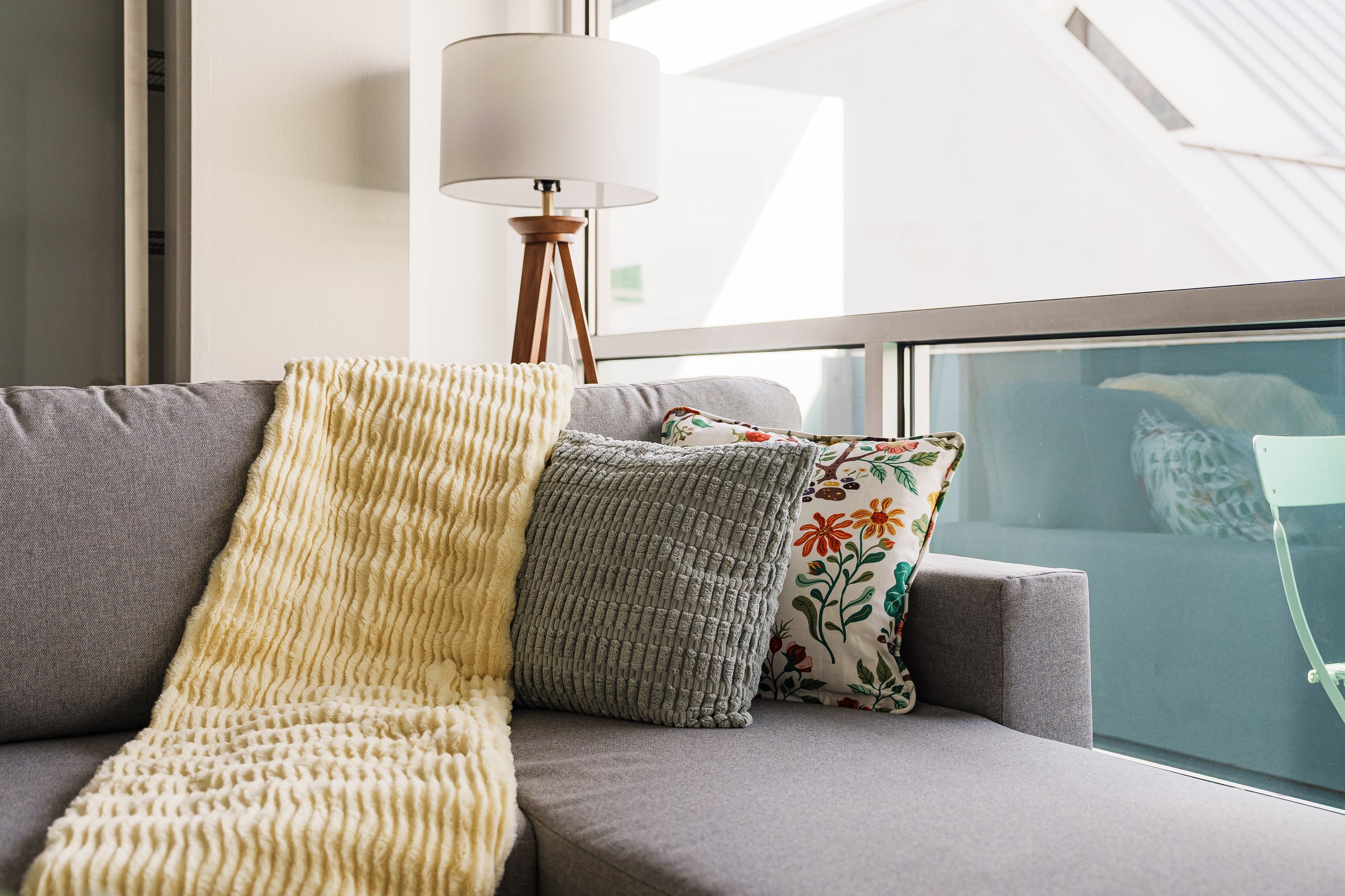 A gray sofa with three decorative pillows and a yellow blanket, next to a window with a modern lamp in the background.