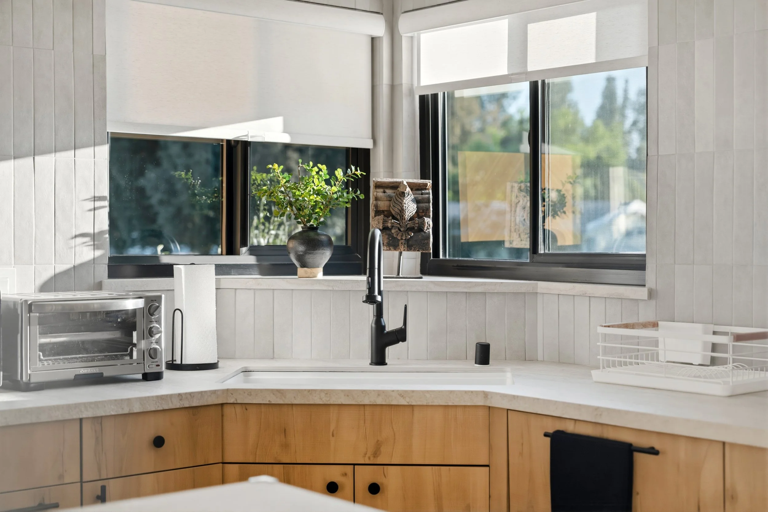 A kitchen scene with a window, potted plant, toaster oven, paper towel holder, and dish rack on a white countertop, with light-colored cabinets below.