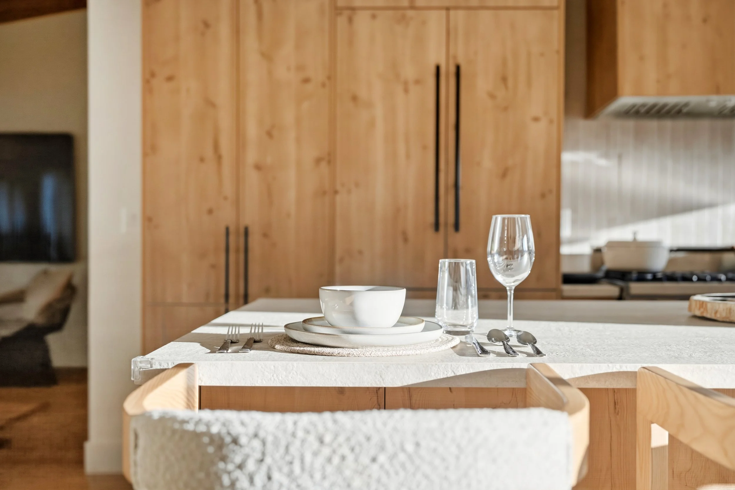 A dining table set with white dinnerware, cutlery, a water glass, and a wine glass in a modern kitchen with wooden cabinets.