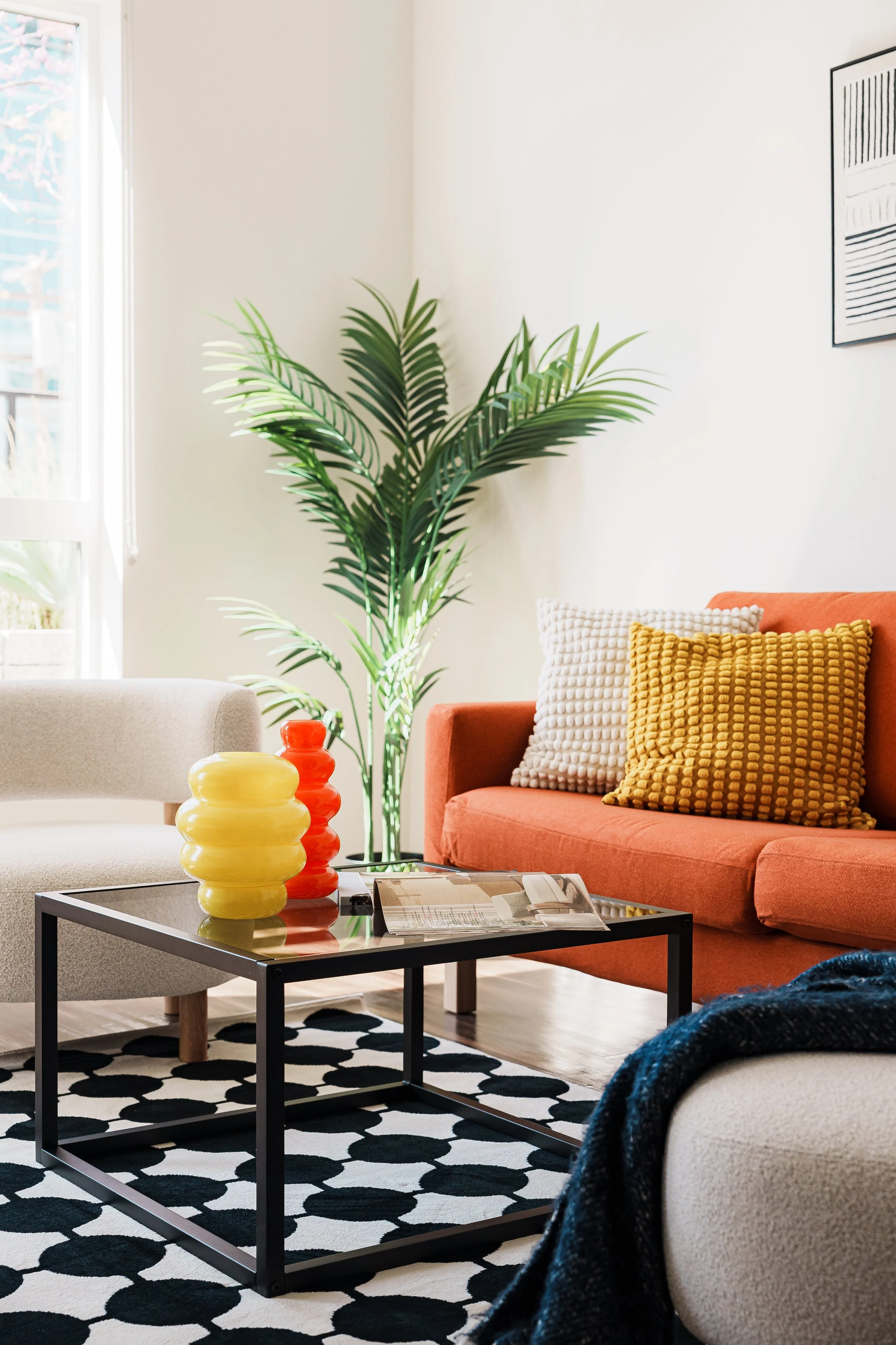 Living room with a large green potted plant, orange sofa with white and yellow textured pillows, black metal coffee table with yellow and orange vases, patterned black and white rug, and a throw blanket.