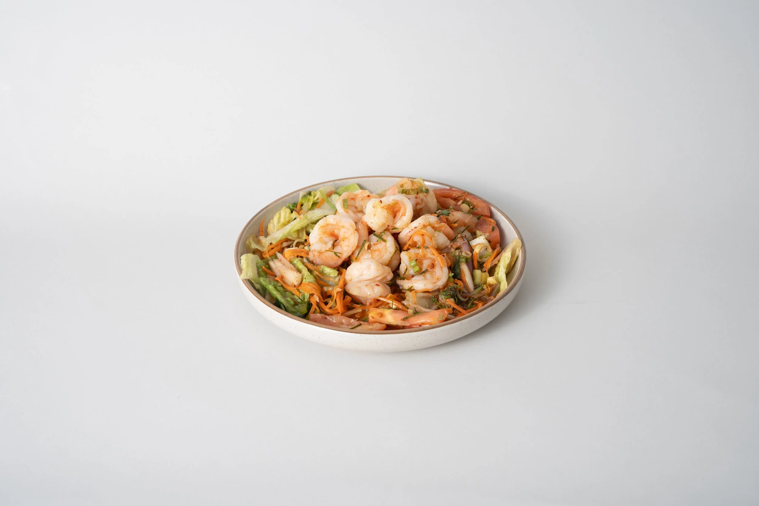 Shrimp salad in a white bowl with lettuce, carrots, and onions on a white surface.