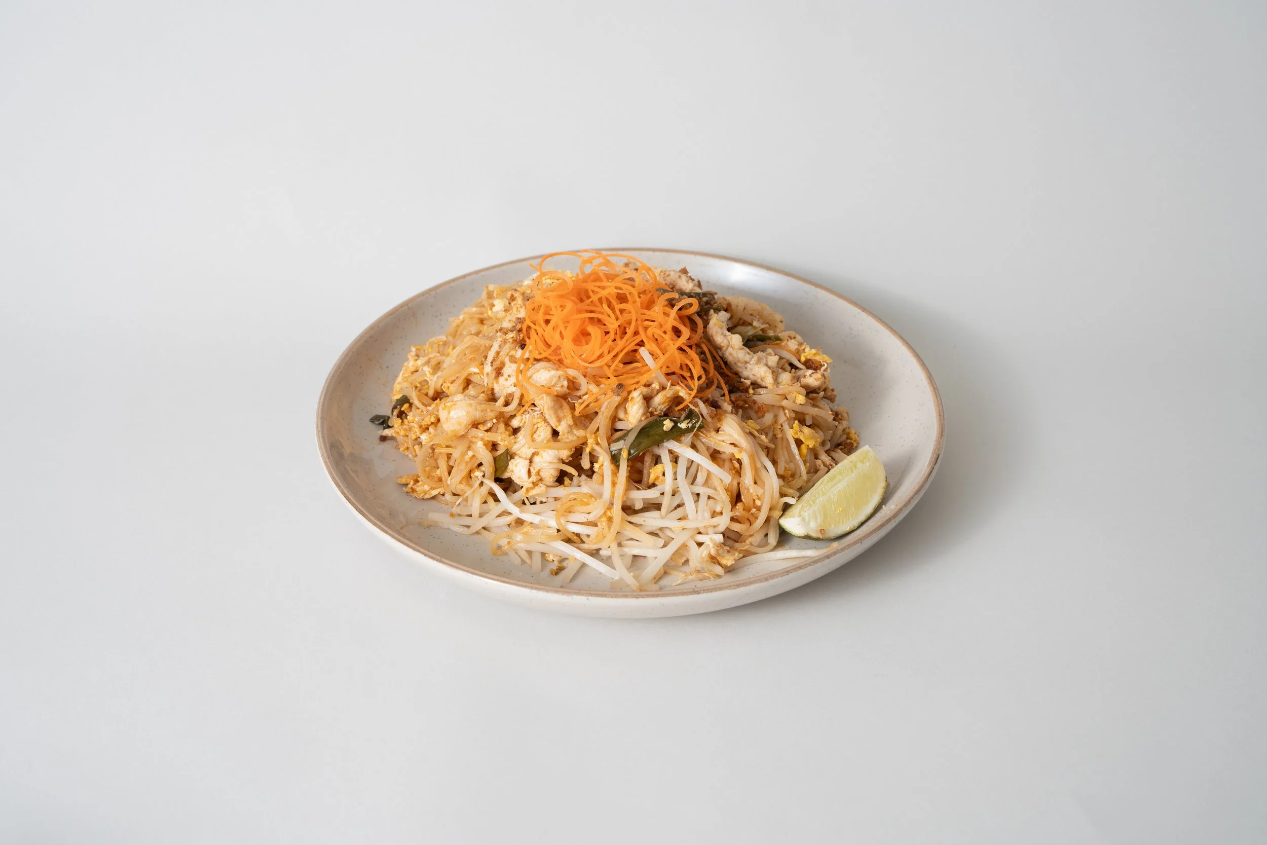 Plate of Pad Thai noodles topped with shredded carrots and lime wedge on white background.