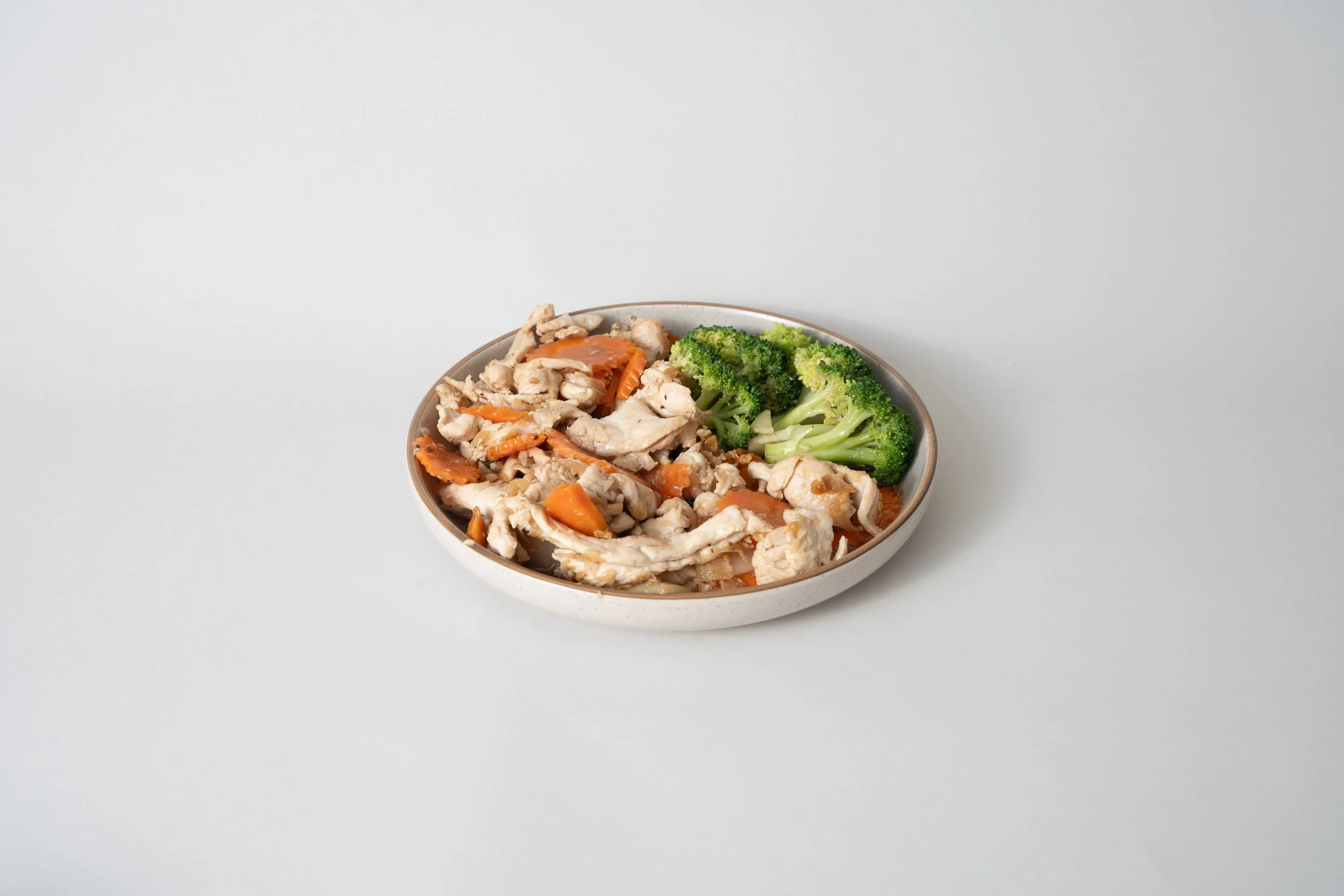 A bowl of cooked chicken and carrots with broccoli on a light background.